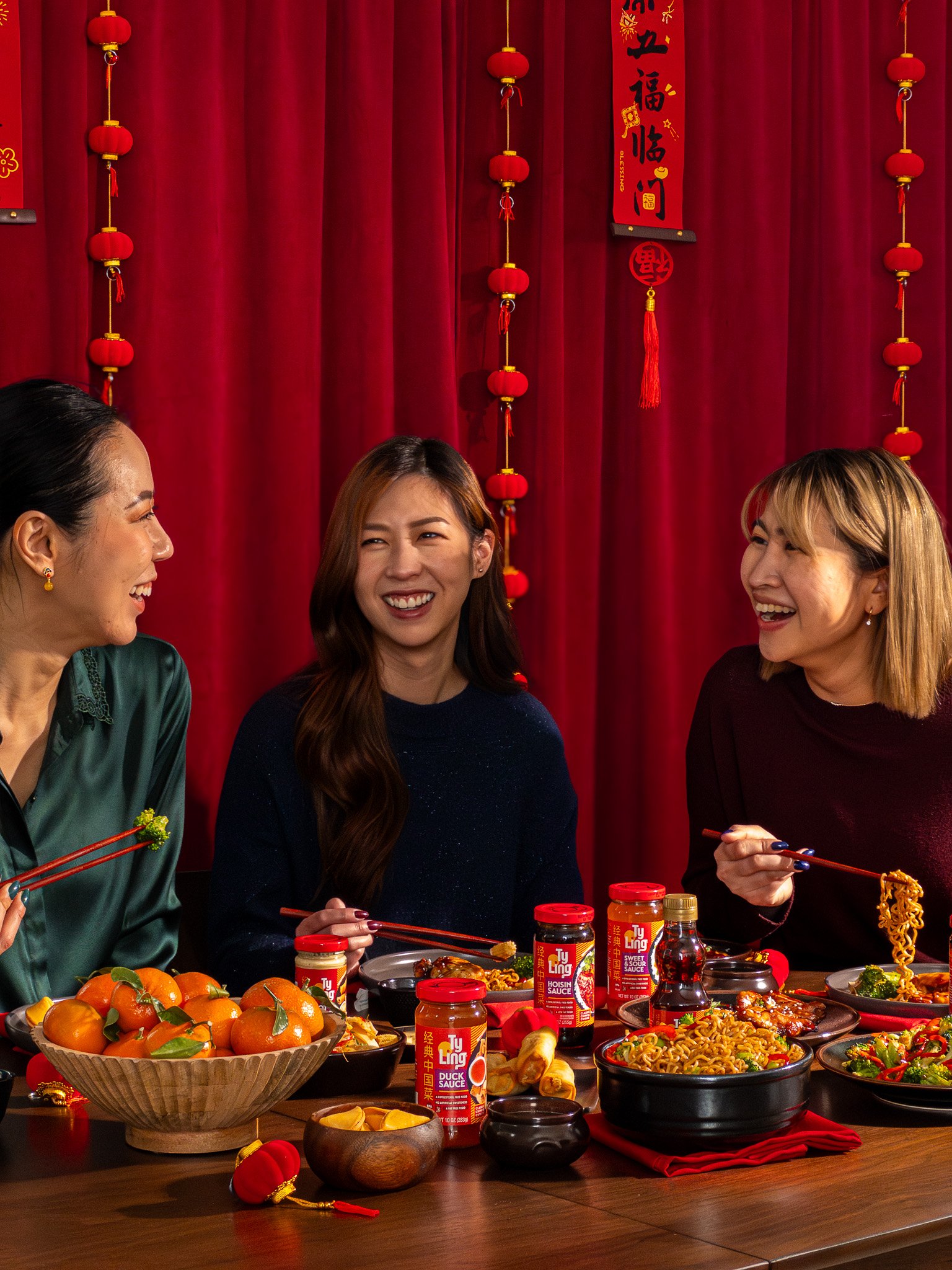 Three women having a joyful conversation at a table filled with Chinese food and condiments, with red lanterns and Chinese decorations hanging on a red curtain background.