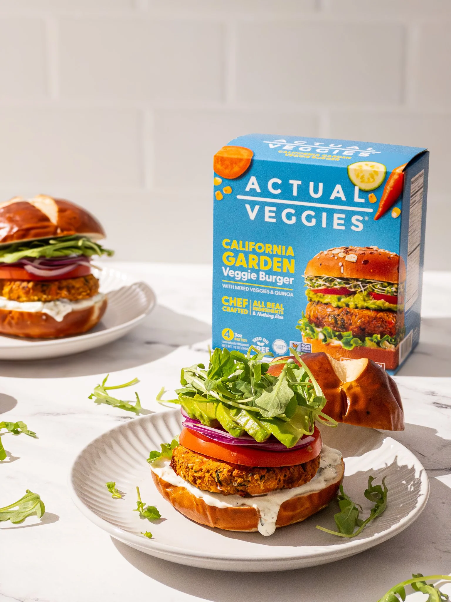 A prepared veggie burger on a white plate with a side of green arugula. Behind it, a blue box of Actual Veggies California Garden Veggie Burger with images of veggies and burger ingredients. The burger includes lettuce, tomato, onion, and a veggie pa