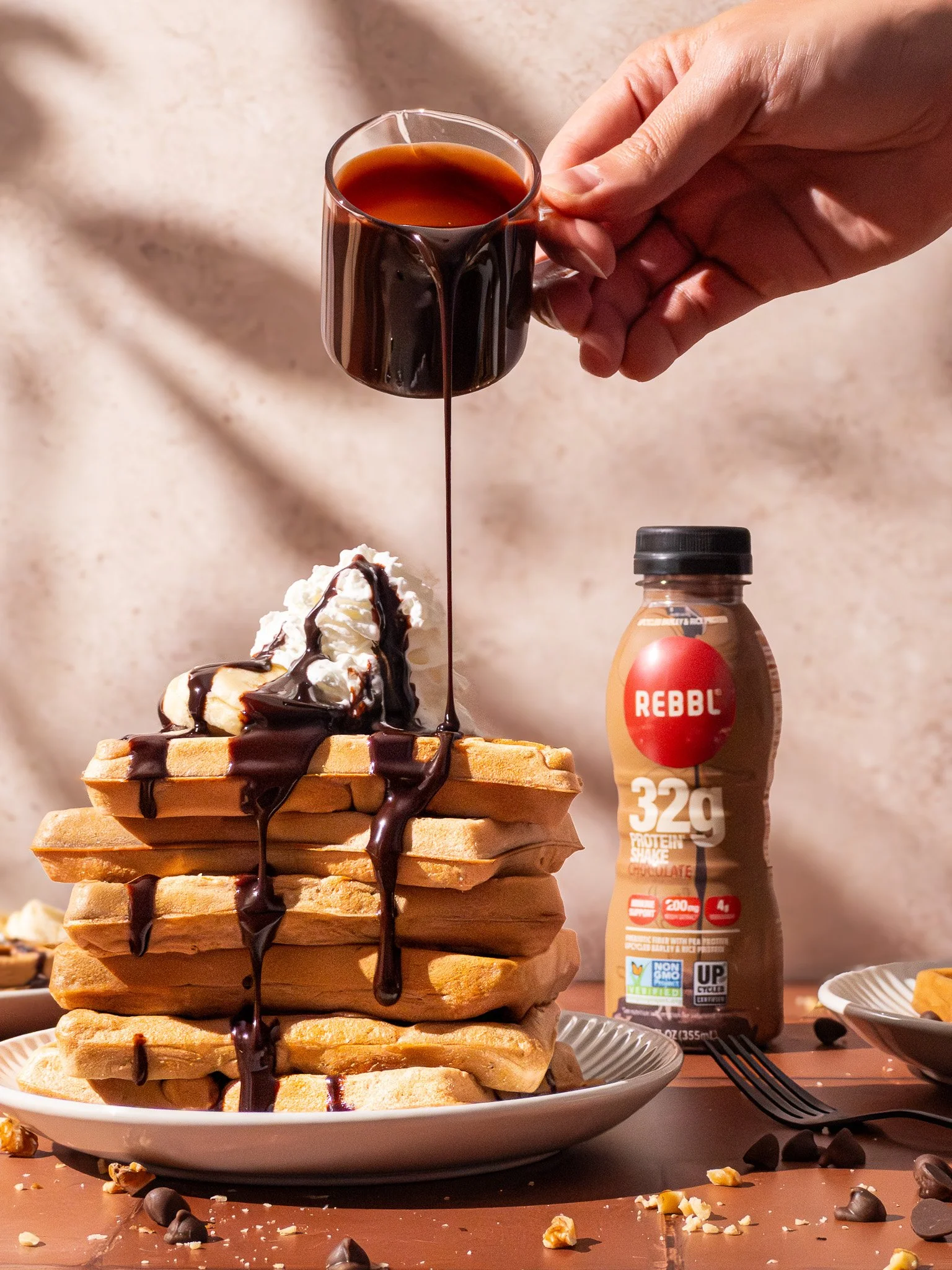 A stack of waffles topped with whipped cream and drizzled with chocolate syrup, with a hand pouring more chocolate syrup from a small glass cup.