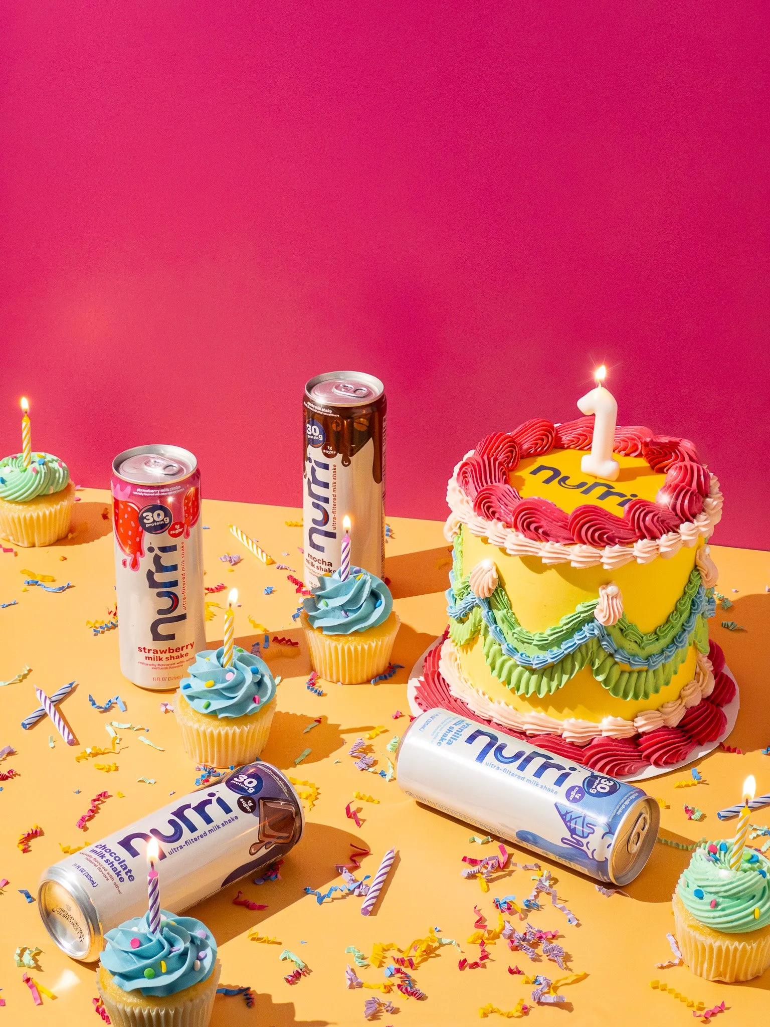 Colorful birthday celebration with cupcakes, a decorated cake with a lit candle, Nuur protein drinks, ribbons, and confetti on a yellow surface with a pink background.