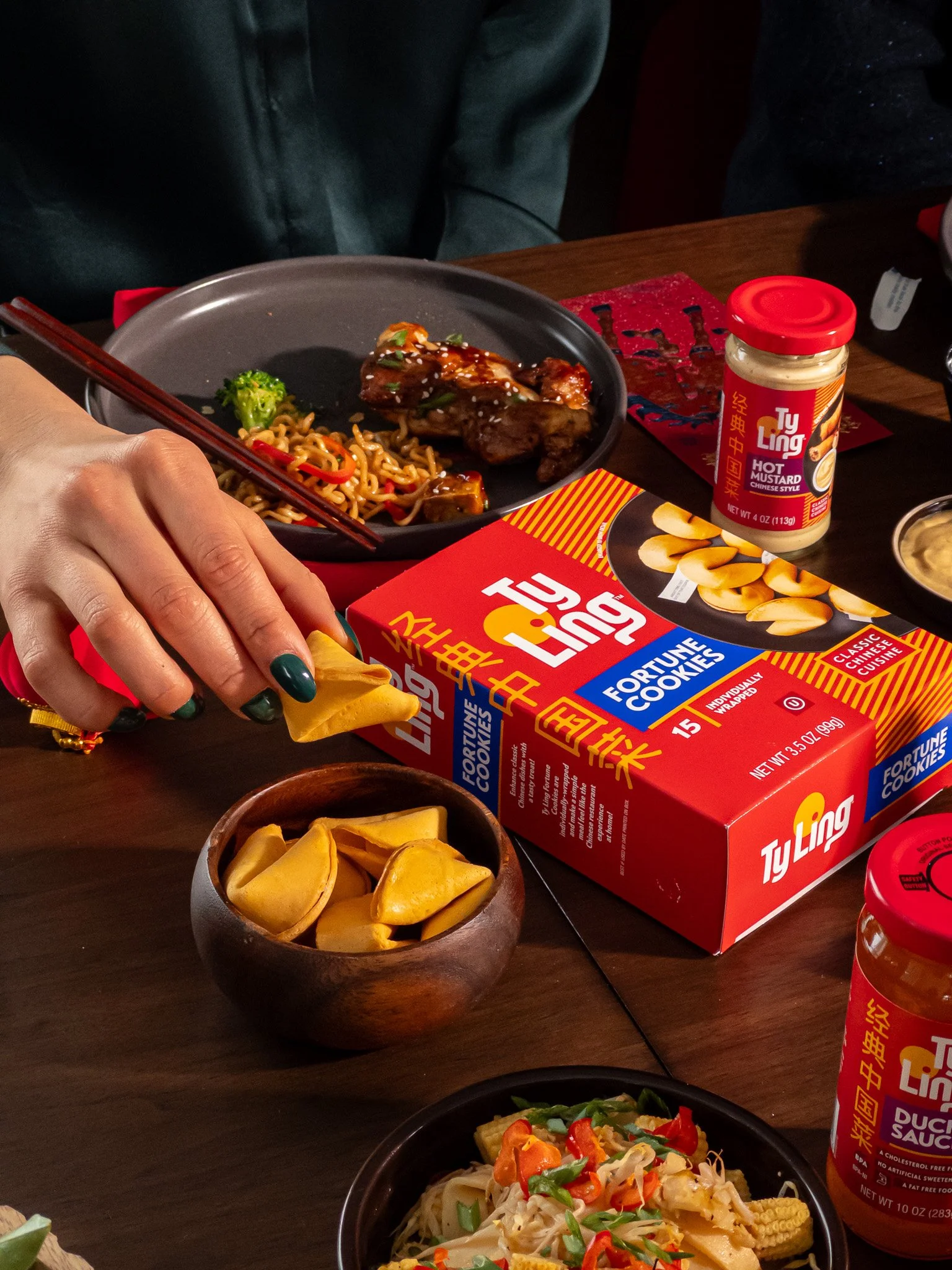 A table with various Asian dishes, including a plate of stir-fried noodles with broccoli and meat, a person removing a fortune cookie from a wooden bowl, boxes of Fortune Cookies, and bottles of Chinese-style sauces.