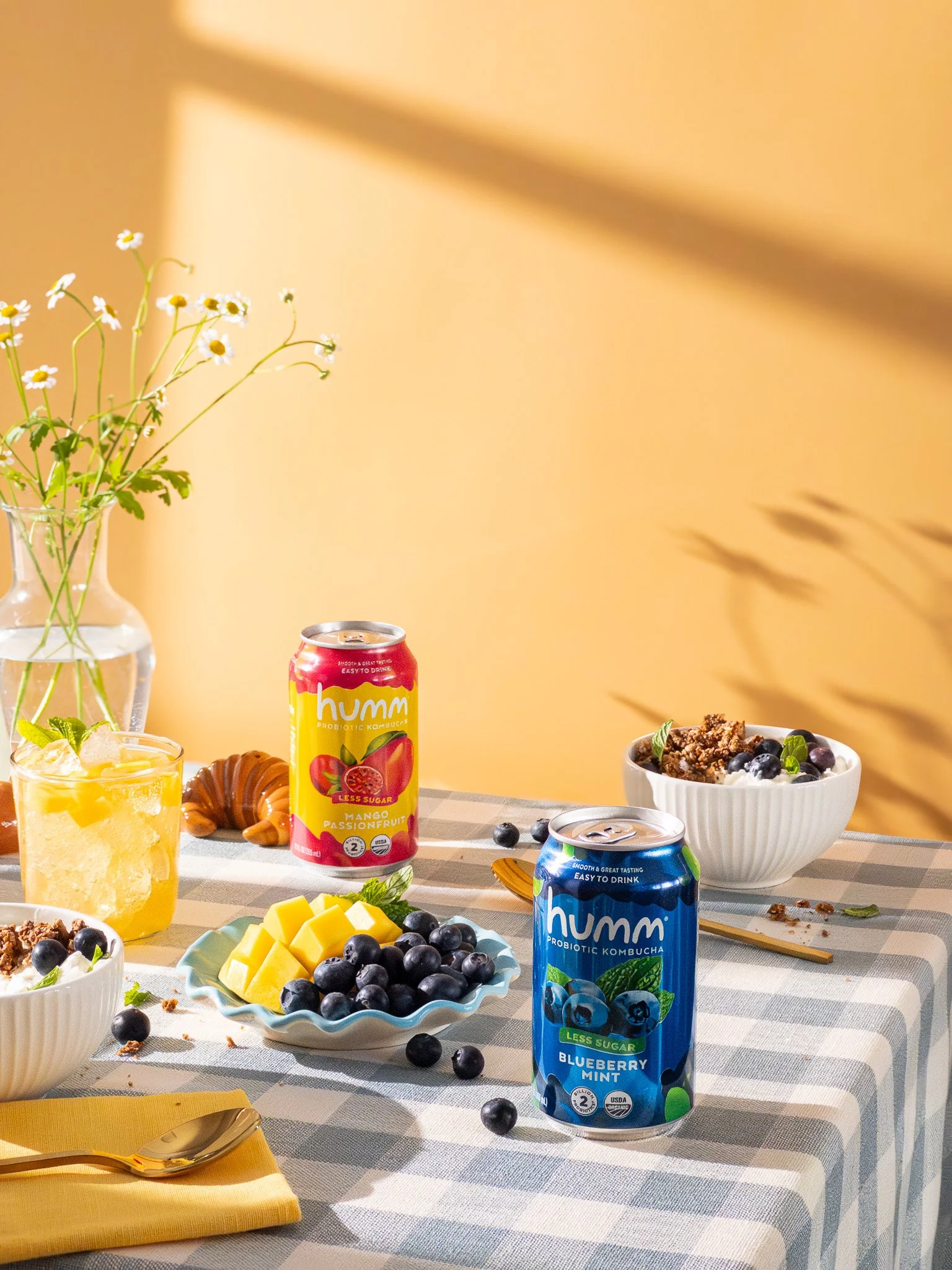 A table with two cans of Humm probiotic kombucha, one blueberry mint flavor and one mango passionfruit flavor, along with bowls of blueberries, yogurt with granola, sliced mango, a glass of lemonade, a croissant, a spoon on a yellow napkin, and a gla