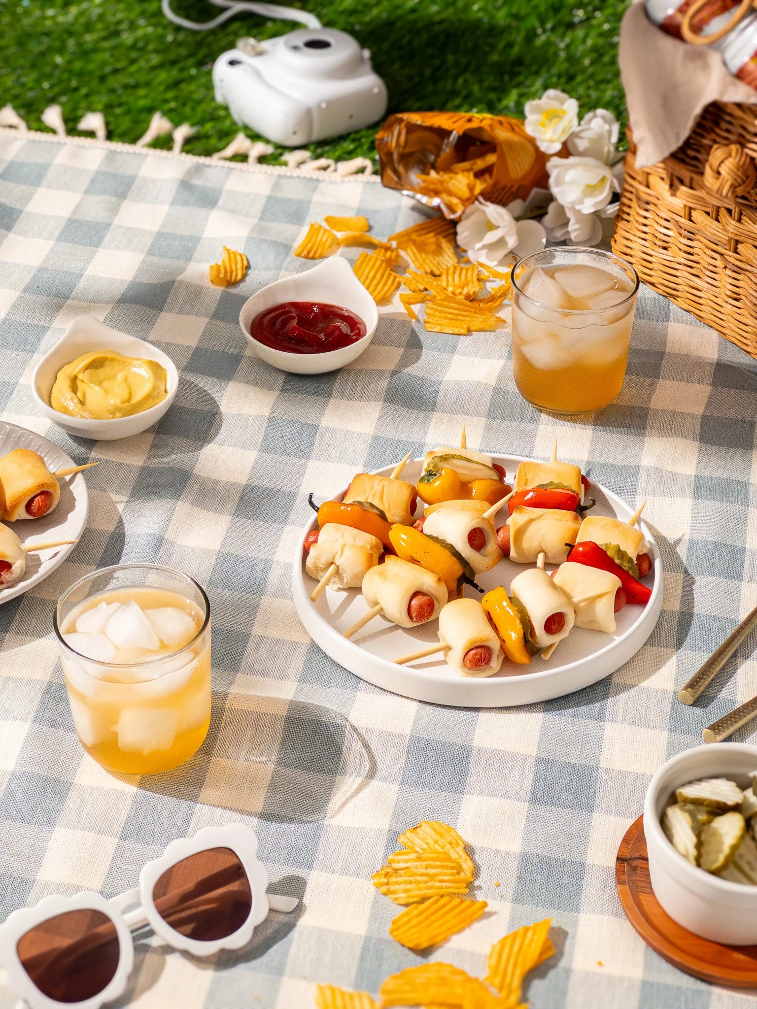 A picnic table with a checkered tablecloth holds a plate of mini hot dogs wrapped in melted cheese and skewered with toothpicks, cups of iced tea, bowls of mustard and ketchup, a basket of chips with some spilled onto the table, a bowl of pickles, a 
