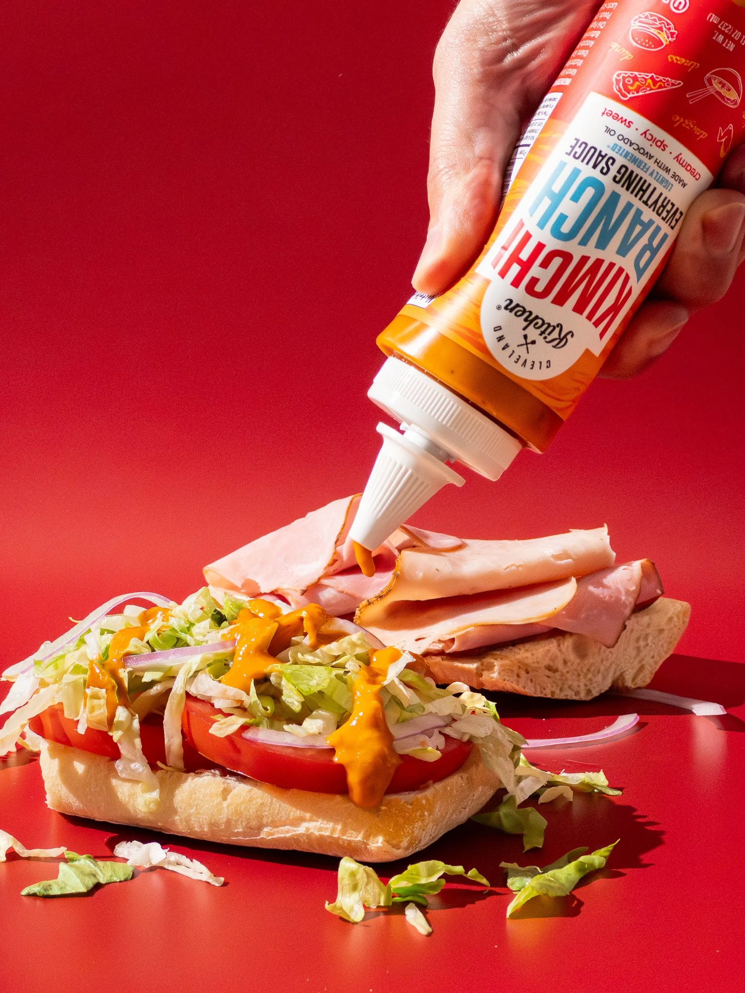 A sandwich being topped with a squeeze of creamy dressing from a ketchup bottle, on a red background.