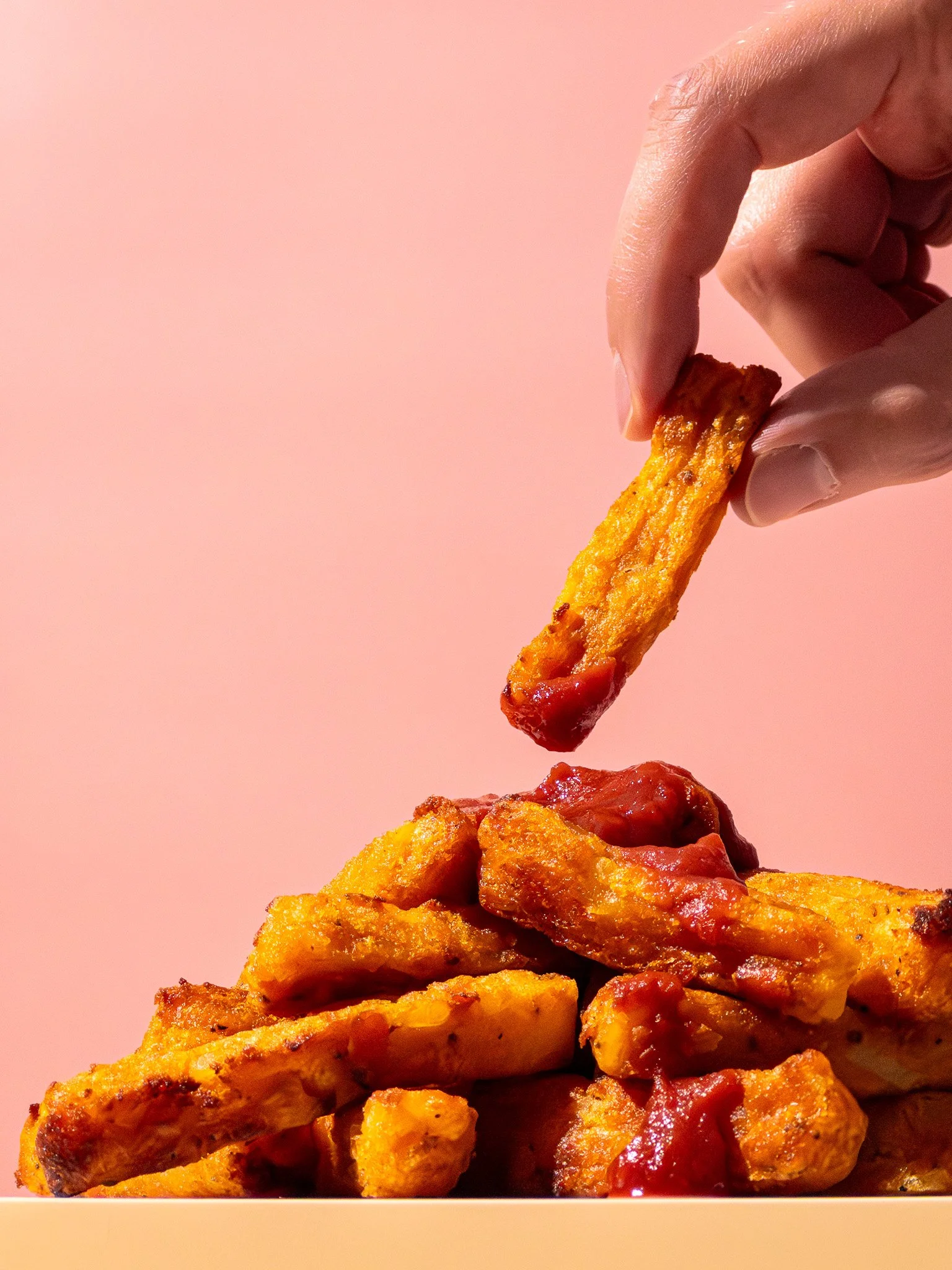 A hand dipping a piece of crispy fried chicken into a pile of fried chicken pieces with ketchup on a pink background.