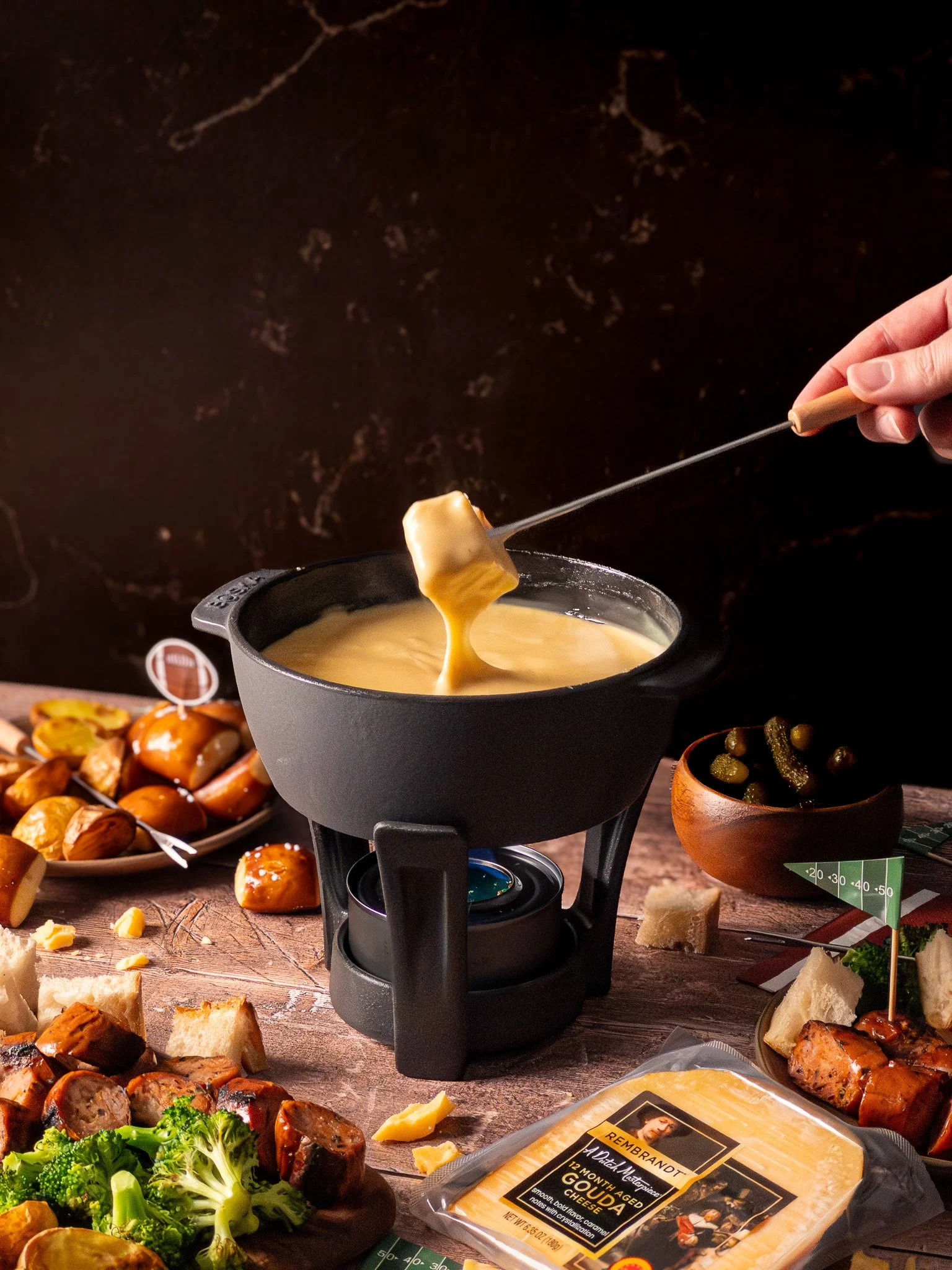 A cheese fondue pot with melted cheese on a wooden table surrounded by cheese, bread, vegetables, and meats.