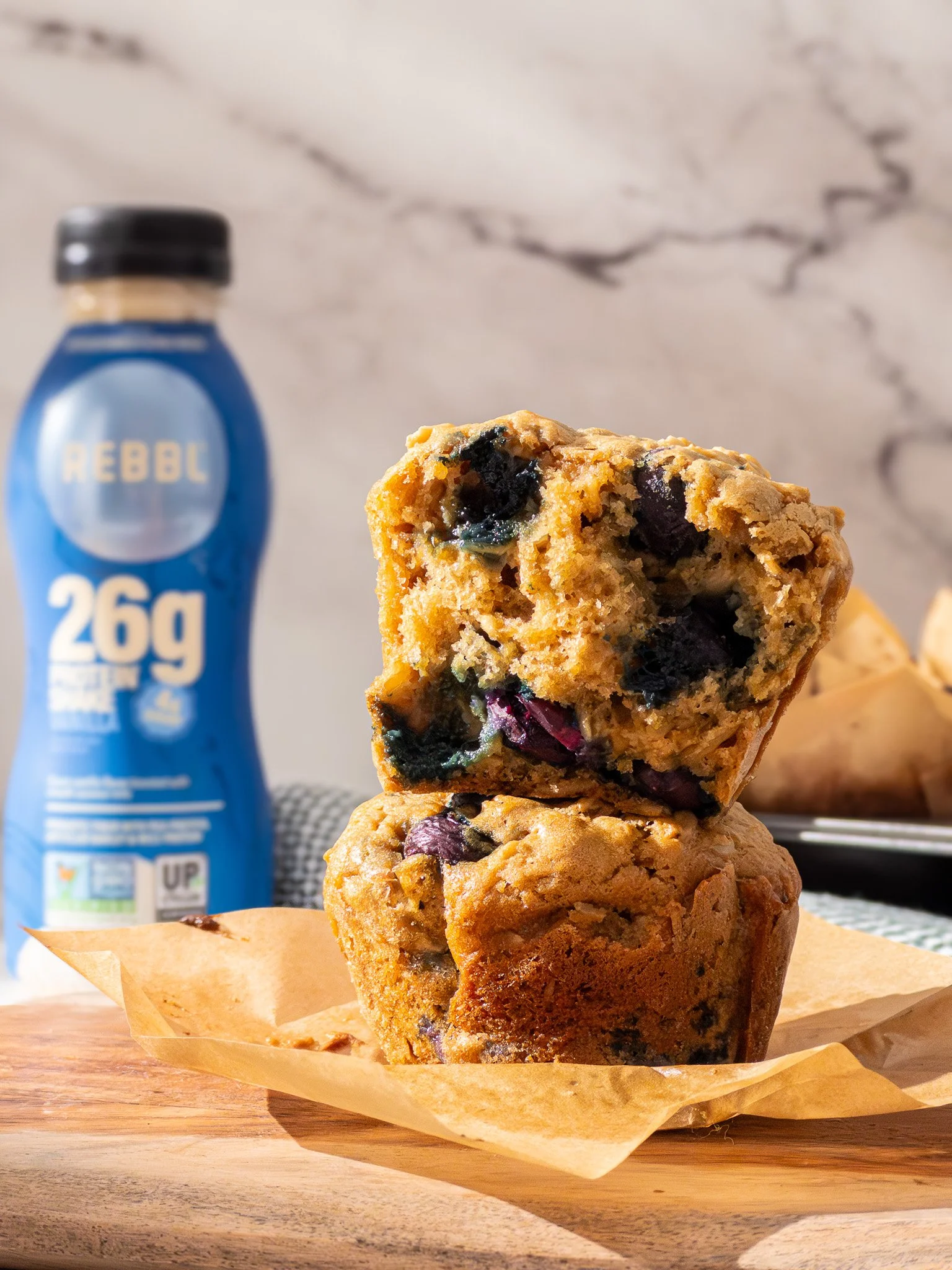 A close-up of a blueberry muffin, one showing the inside with blueberries, on a piece of parchment paper on a wooden surface, with a bottle of Rebbl plant-based protein drink in the background.