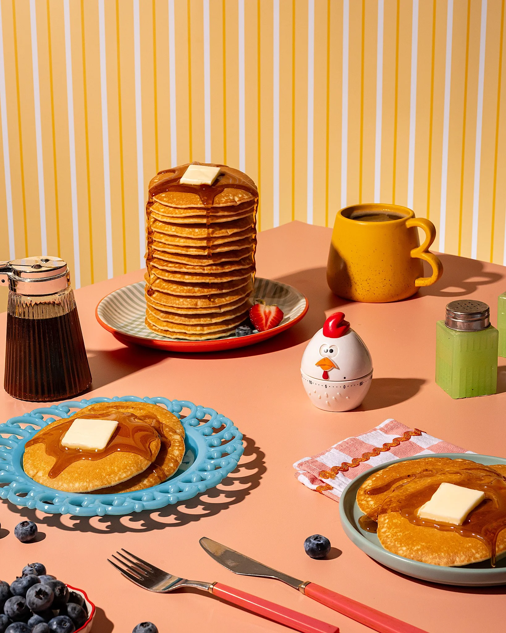 Stack of pancakes with butter and syrup, blueberry pancakes with butter and syrup, mug of coffee, syrup dispenser, salt and pepper shakers, fruit, plus a chicken-shaped timer, on pink table with yellow and white striped wallpaper.