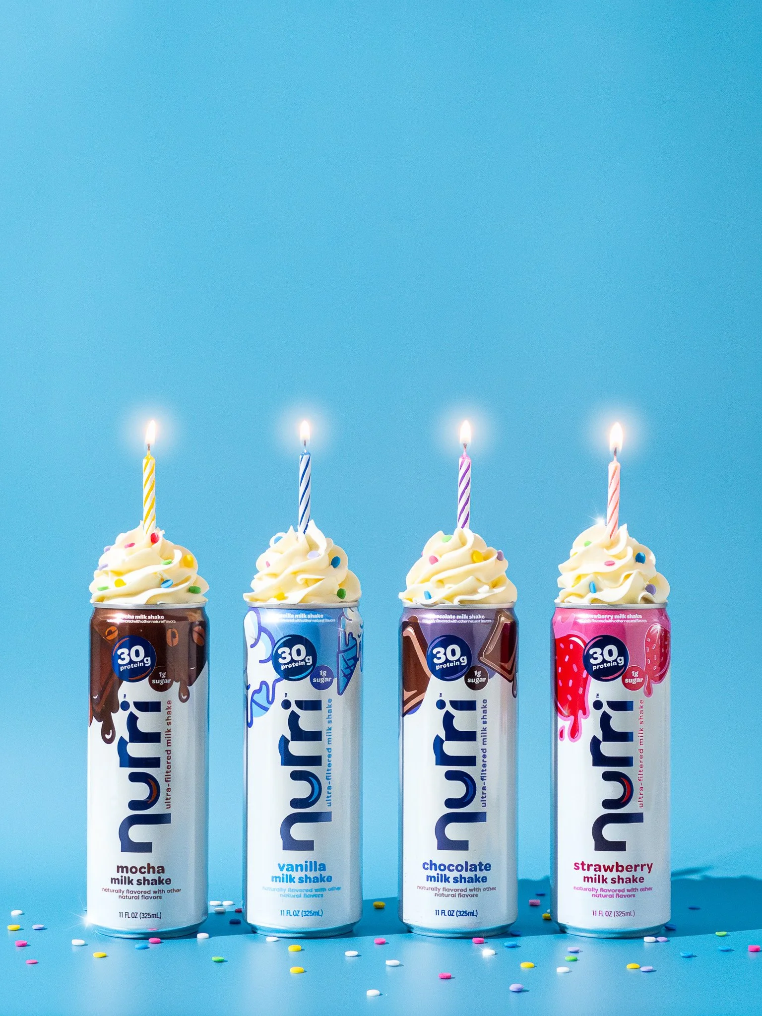 Four cans of flavored milk shake drinks with whipped cream and candles on top, decorated with colorful sprinkles, against a blue background.