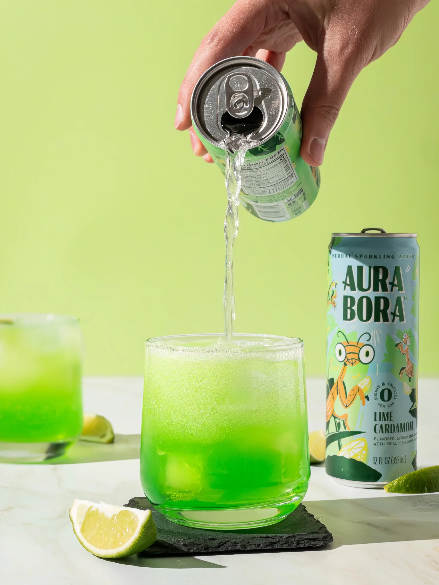 A hand pouring sparkling water from a can into a green drink on a marble surface with lime wedges and a can of Aura Bora Lime Cardamom flavored sparkling water visible in the background.