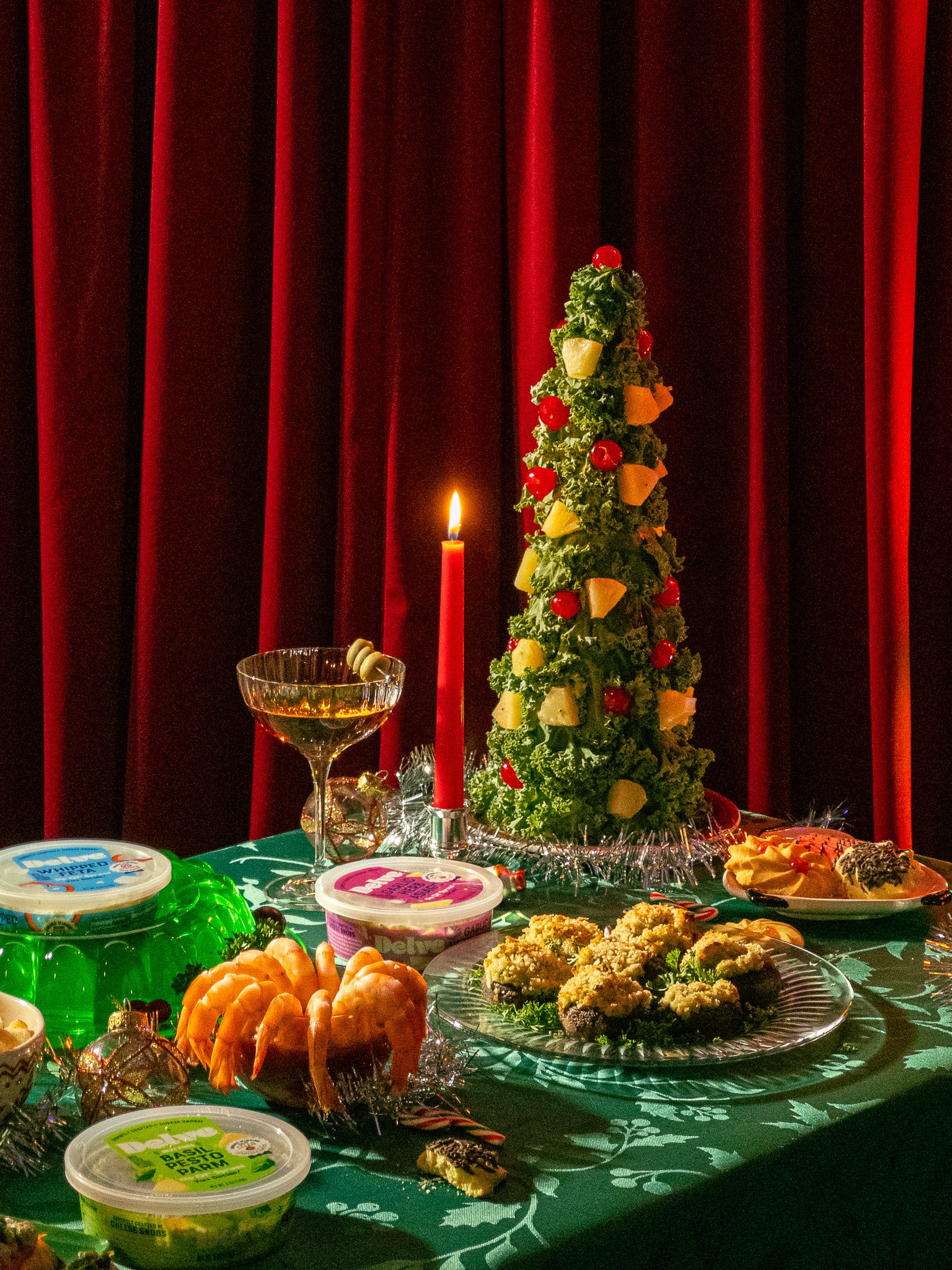 Christmas table with a small decorated tree, a red candle, and various holiday foods and desserts, set against red curtains.