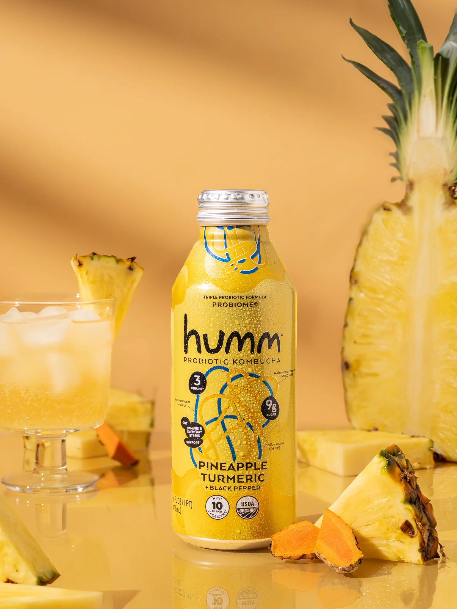 A bottle of HUMM probiotic kombucha with pineapple, turmeric, and black pepper flavor next to pineapple pieces and a glass of pineapple drink, set against a yellow background.