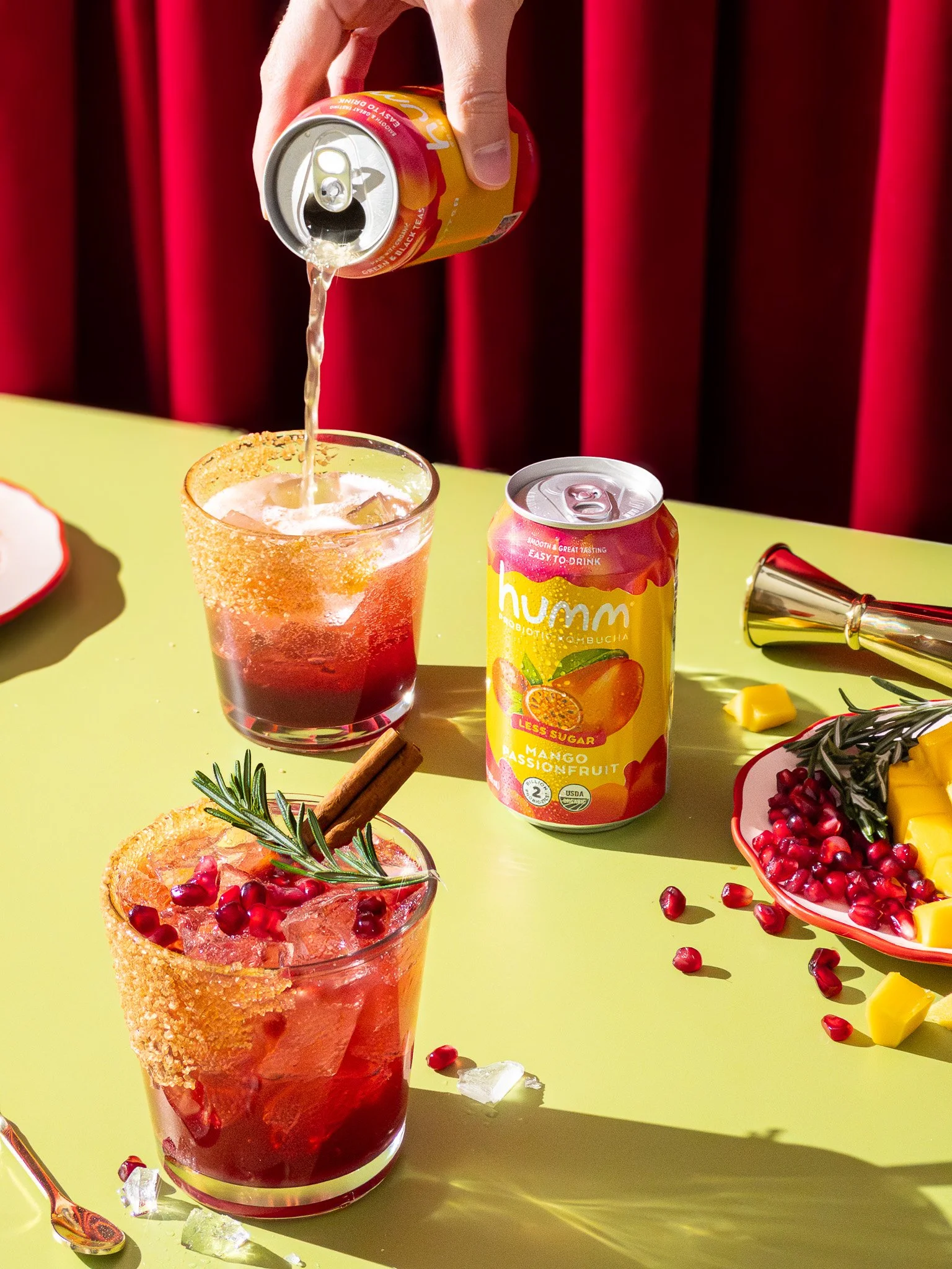 A hand pouring a canned beverage into a glass with ice, surrounded by pomegranate seeds and garnished with a sprig of rosemary and a cinnamon stick, on a yellow table with additional drinks, fruit, and party items.