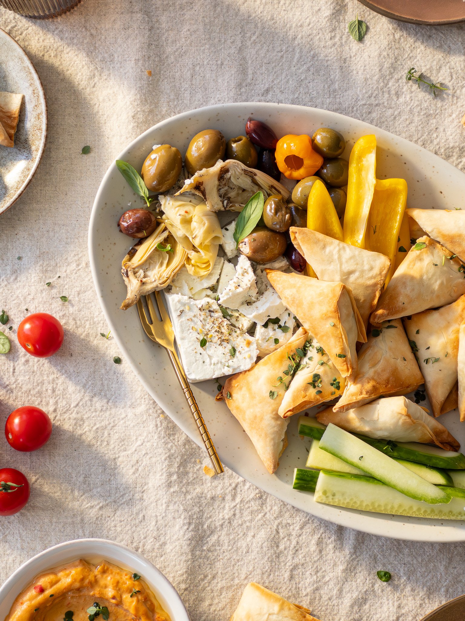 A large white platter with assorted Greek appetizers including olives, feta cheese, stuffed grape leaves, pita triangles, and cucumber slices on a beige tablecloth.