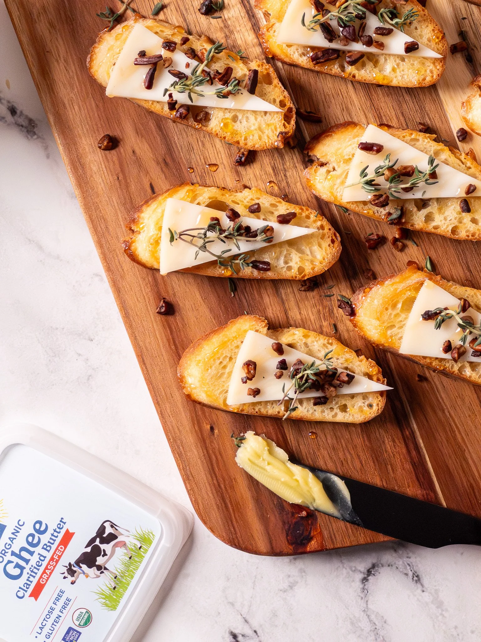 Sliced baguette pieces topped with melted cheese, black garlic chips, and fresh thyme, arranged on a wooden serving board. Butter is being spread on one piece with a small knife, and a container of organic ghee with cow illustrations is nearby.