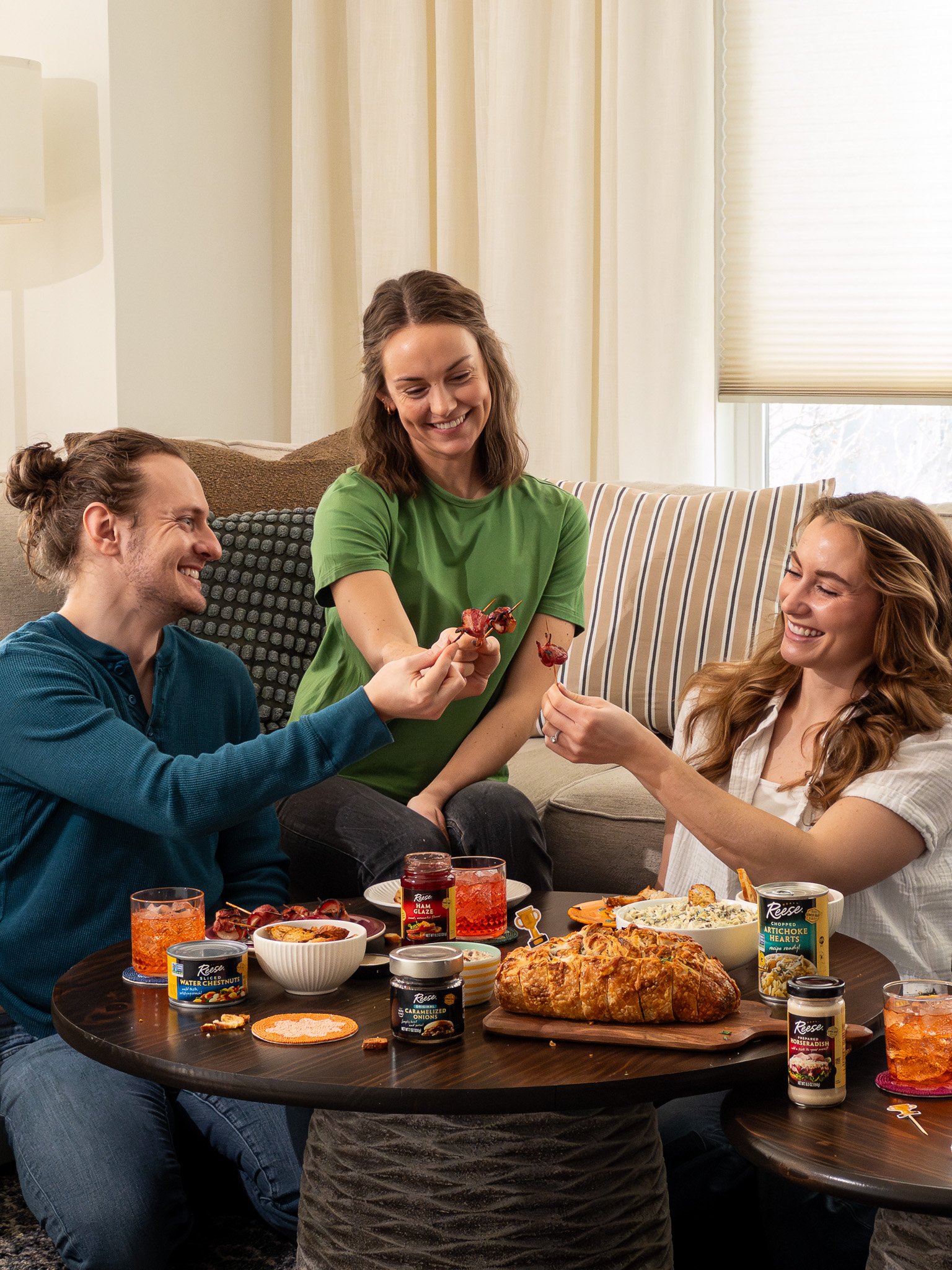 Three friends sitting around a table enjoying a meal, with two of them passing food to each other while smiling and laughing in a cozy living room.