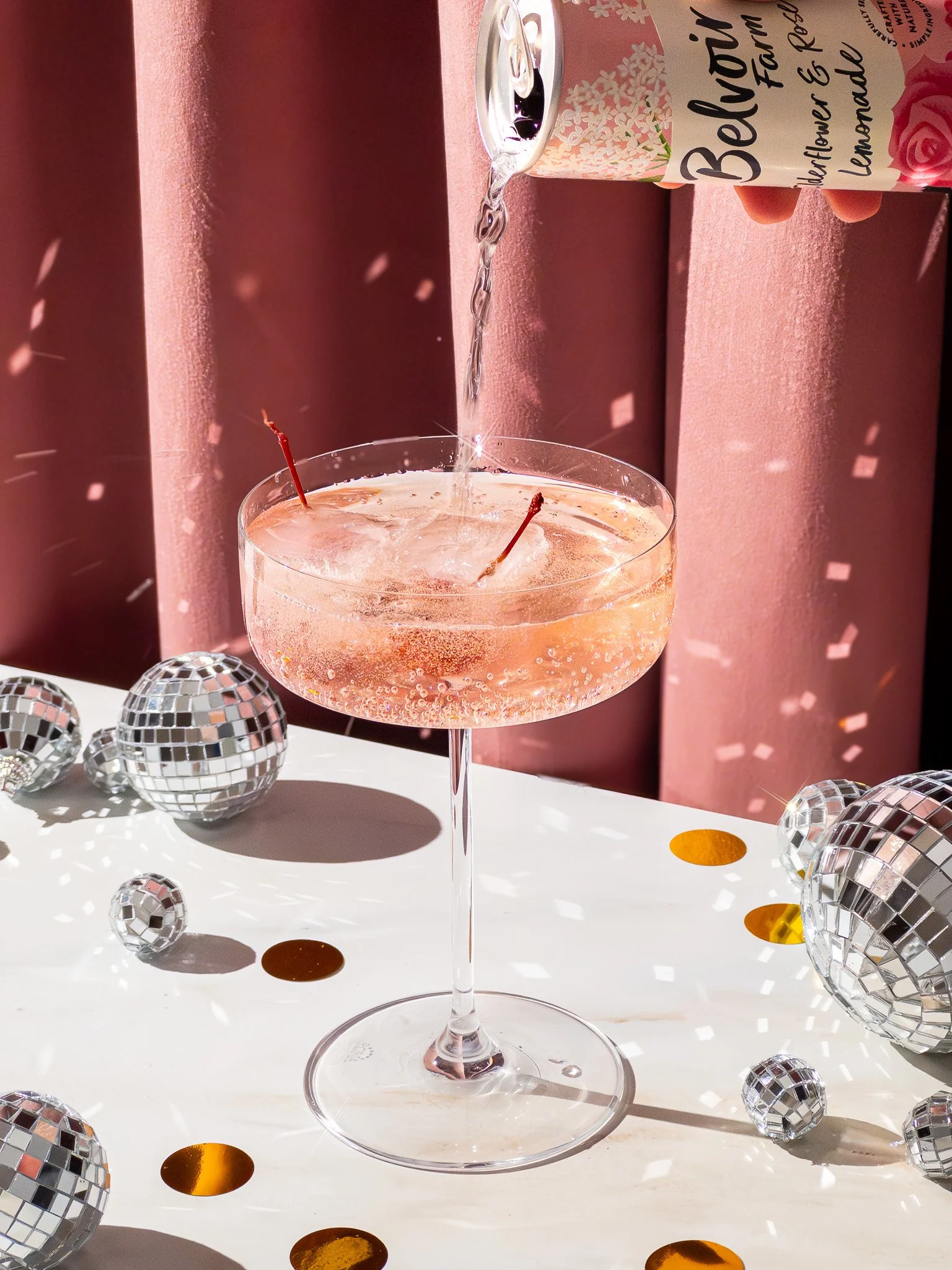 A pink cocktail in a coupe glass with cherries, being poured with sparkling water from a can, surrounded by disco balls and gold confetti on a white table.