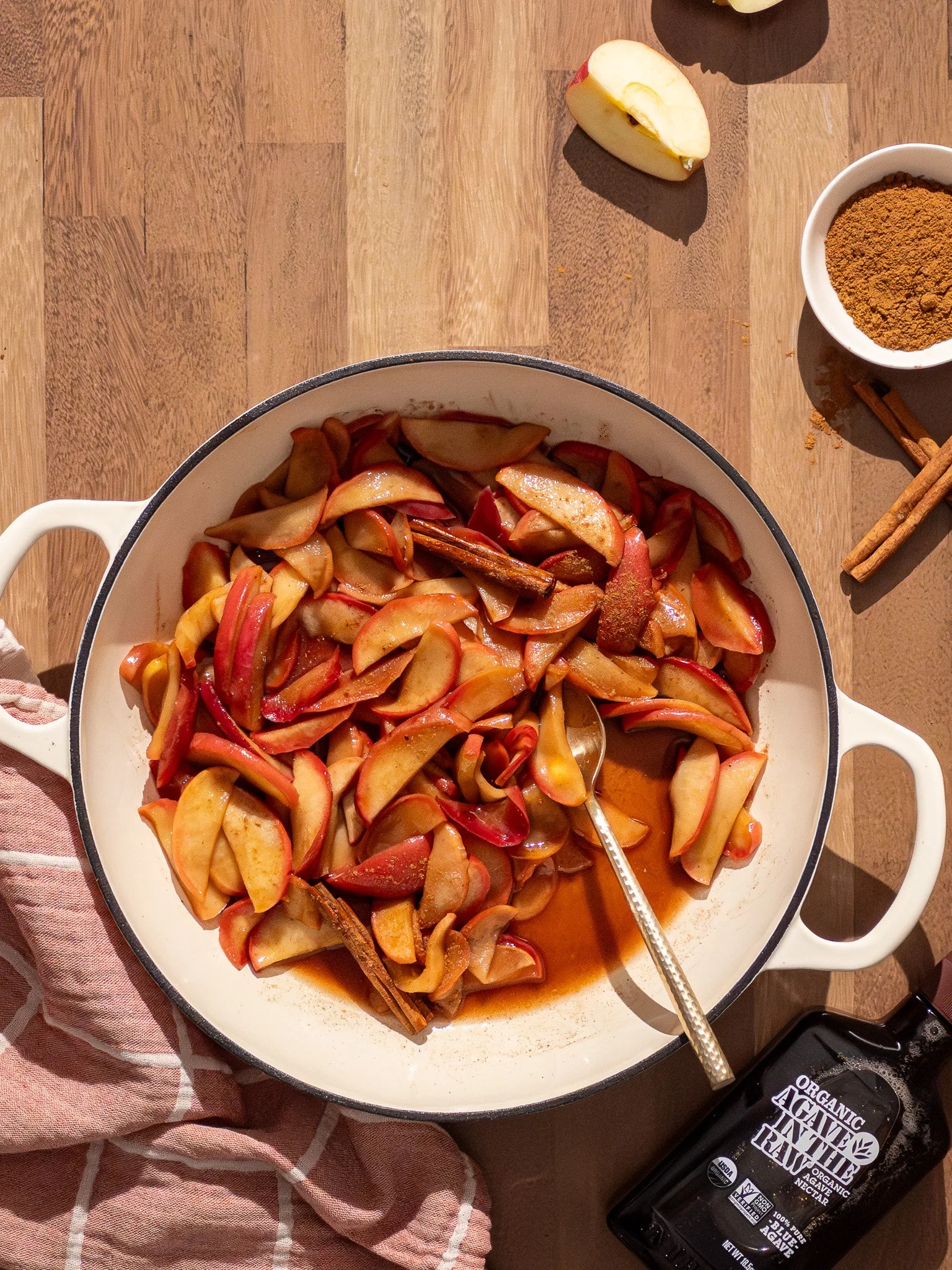 A large white pot filled with cooked apple slices and cinnamon sticks. Surrounding the pot are a halved apple, a small bowl of ground cinnamon, a bottle of organic agave nectar, and cinnamon sticks, all on a wooden surface.