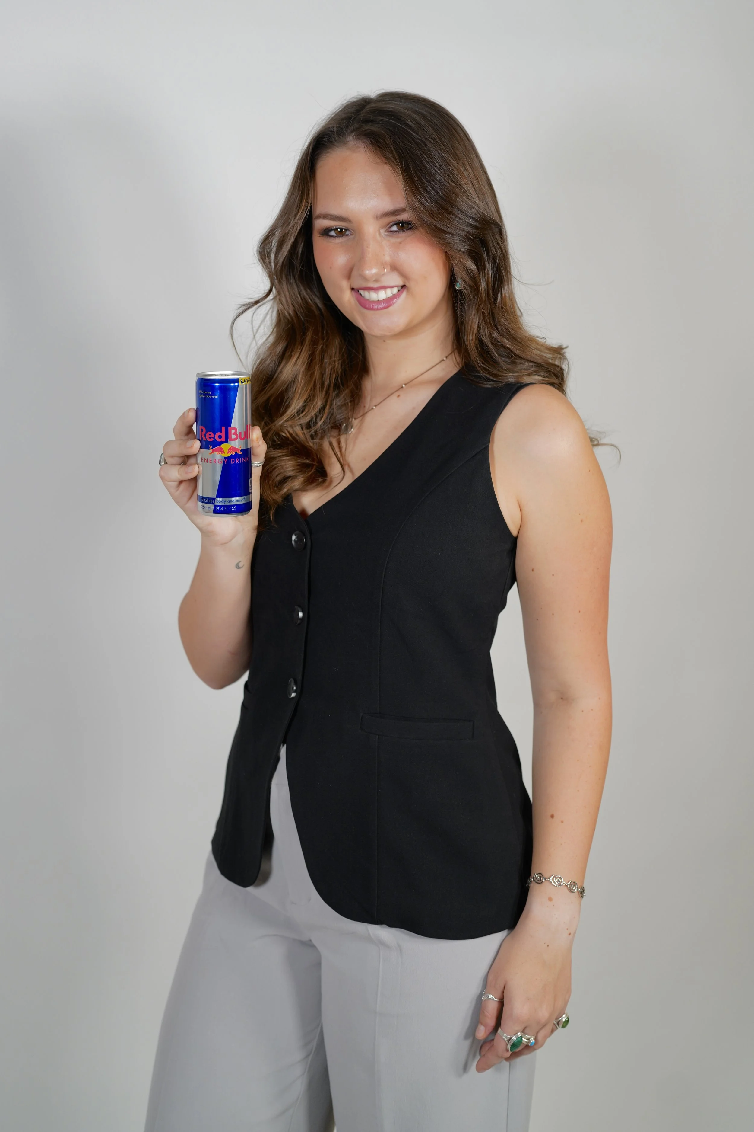 A young woman with long wavy brown hair smiling and holding a can of Red Bull energy drink against a plain white background.