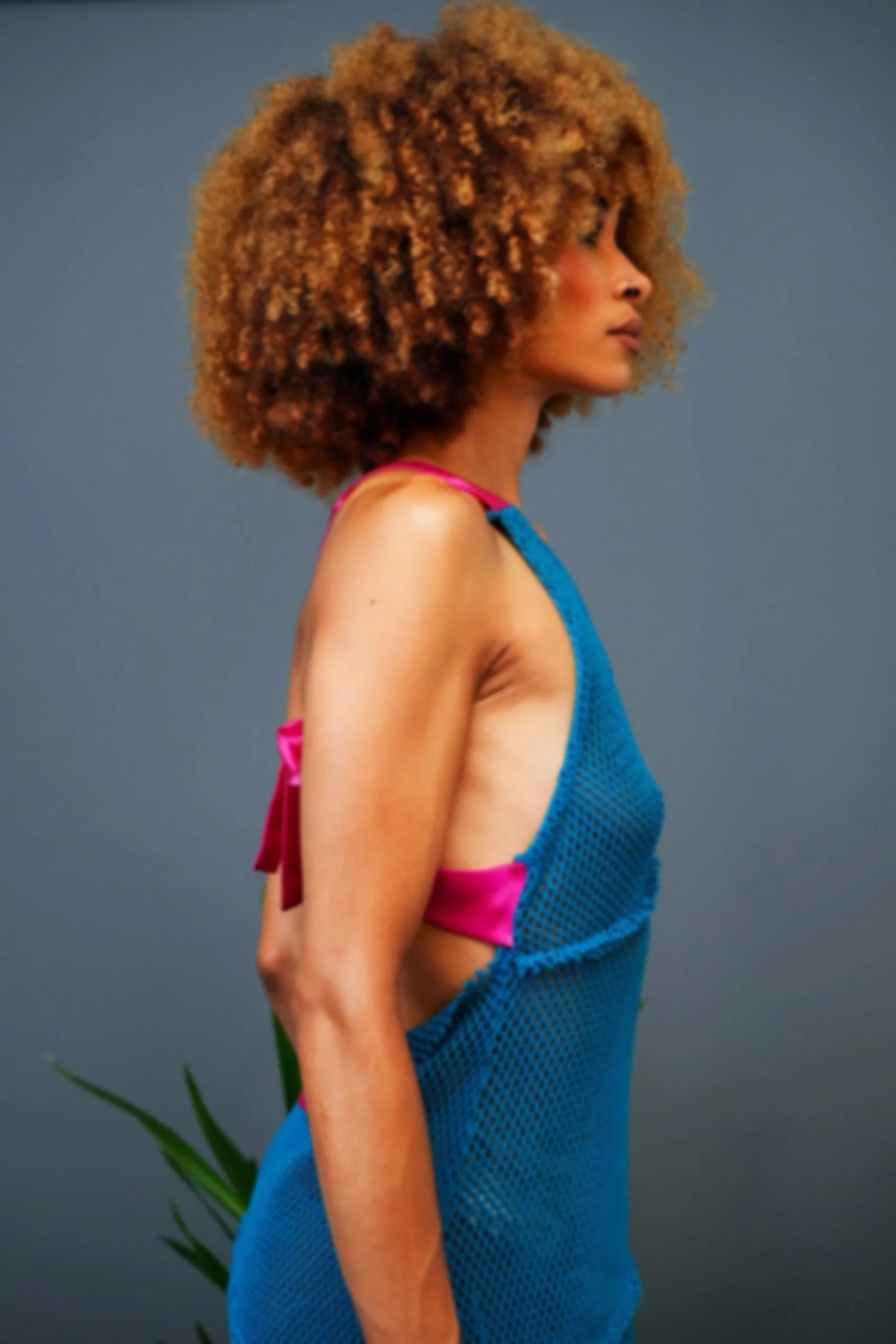 Profile of a woman with curly, voluminous, light brown hair wearing a blue mesh dress with pink straps and a pink bow on the back, standing against a gray background.