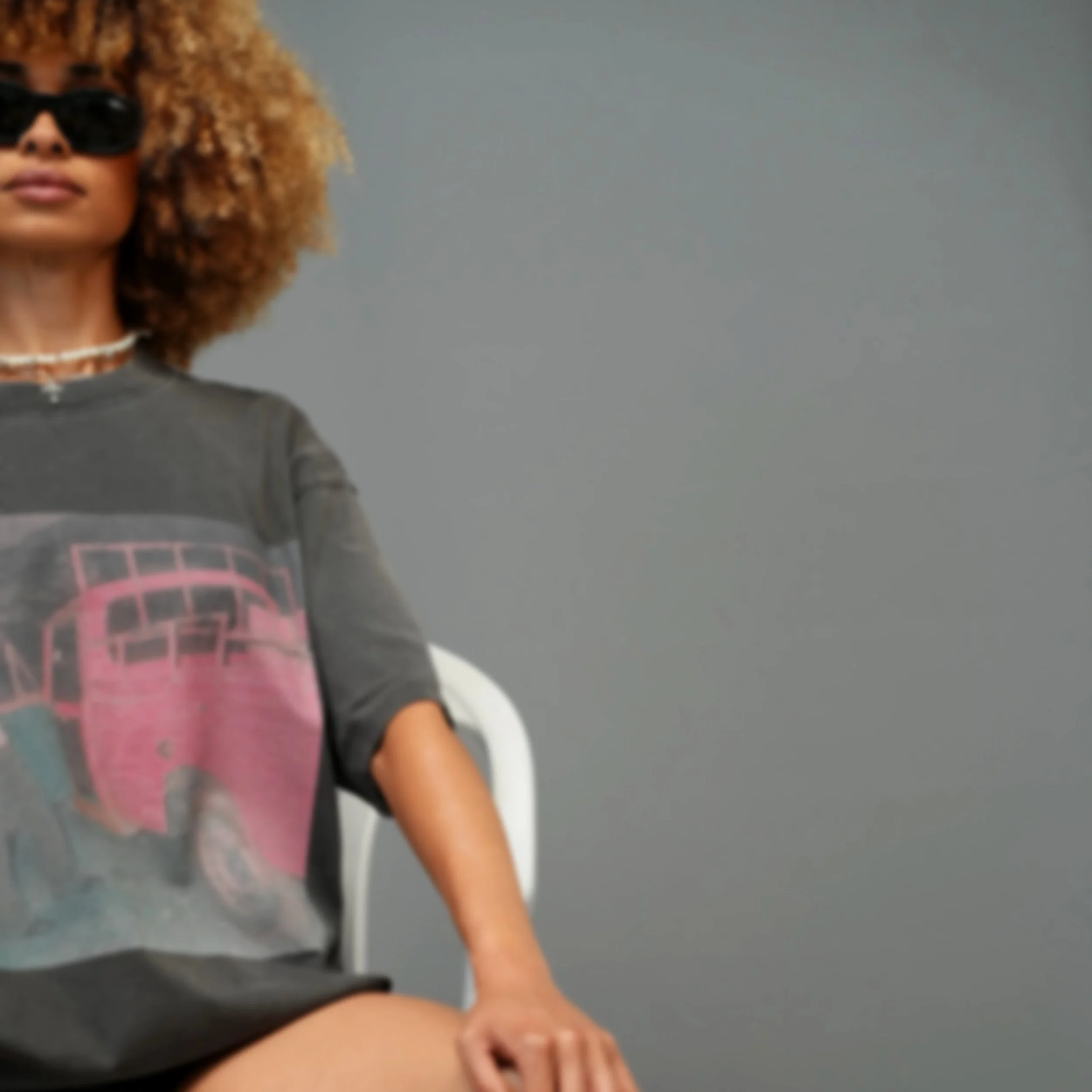 A person with curly blond hair wearing sunglasses and a gray shirt with a pink geometric design, standing against a gray background.