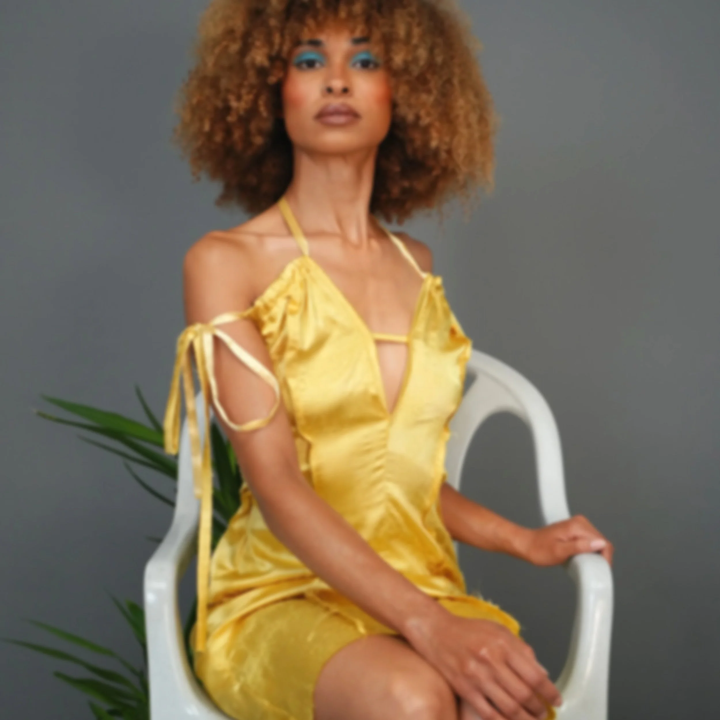 A woman with curly hair wearing a yellow top with straps sitting in a white chair against a gray background.