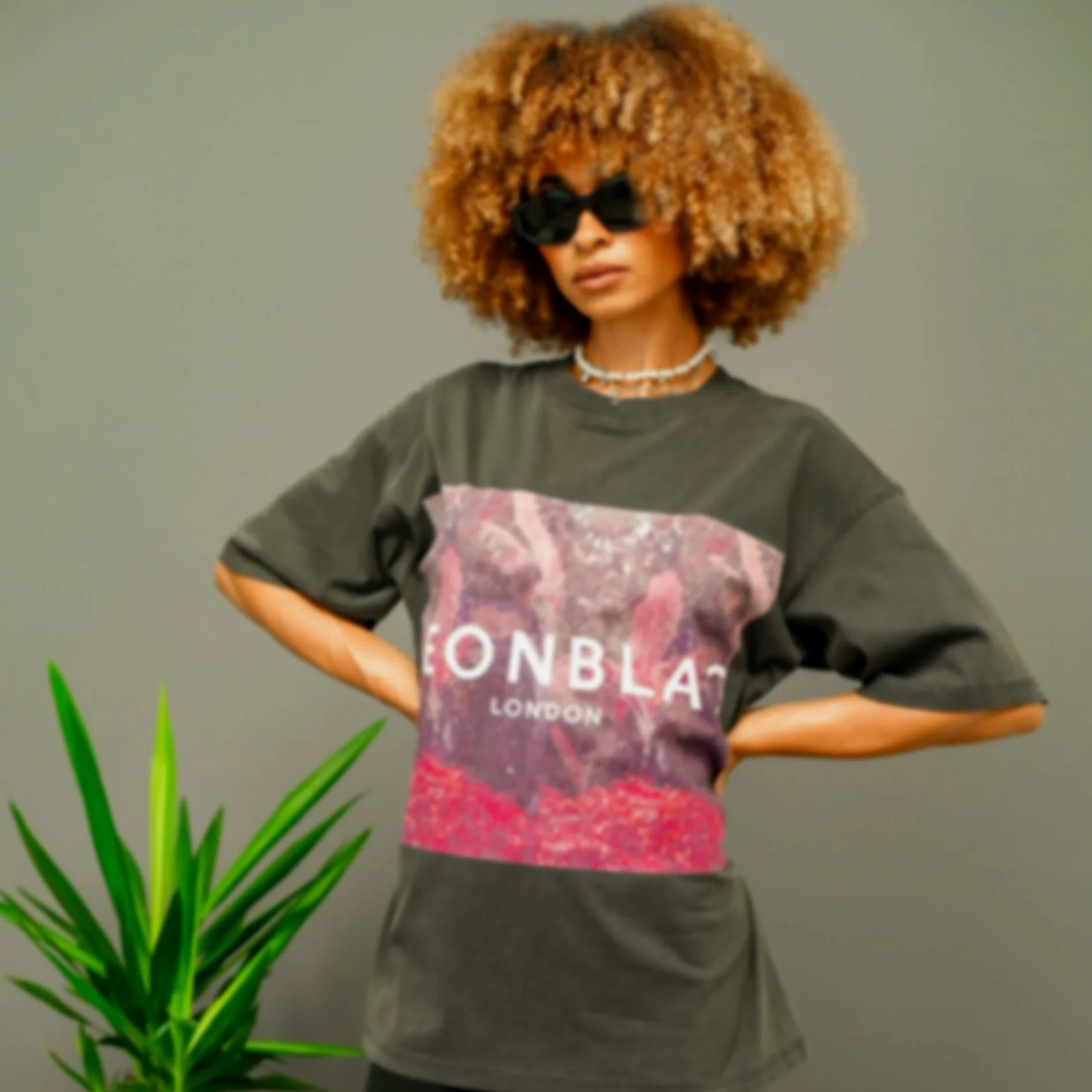 Person with large curly hair wearing dark sunglasses, a graphic t-shirt, and dark pants standing near a green plant against a plain background.