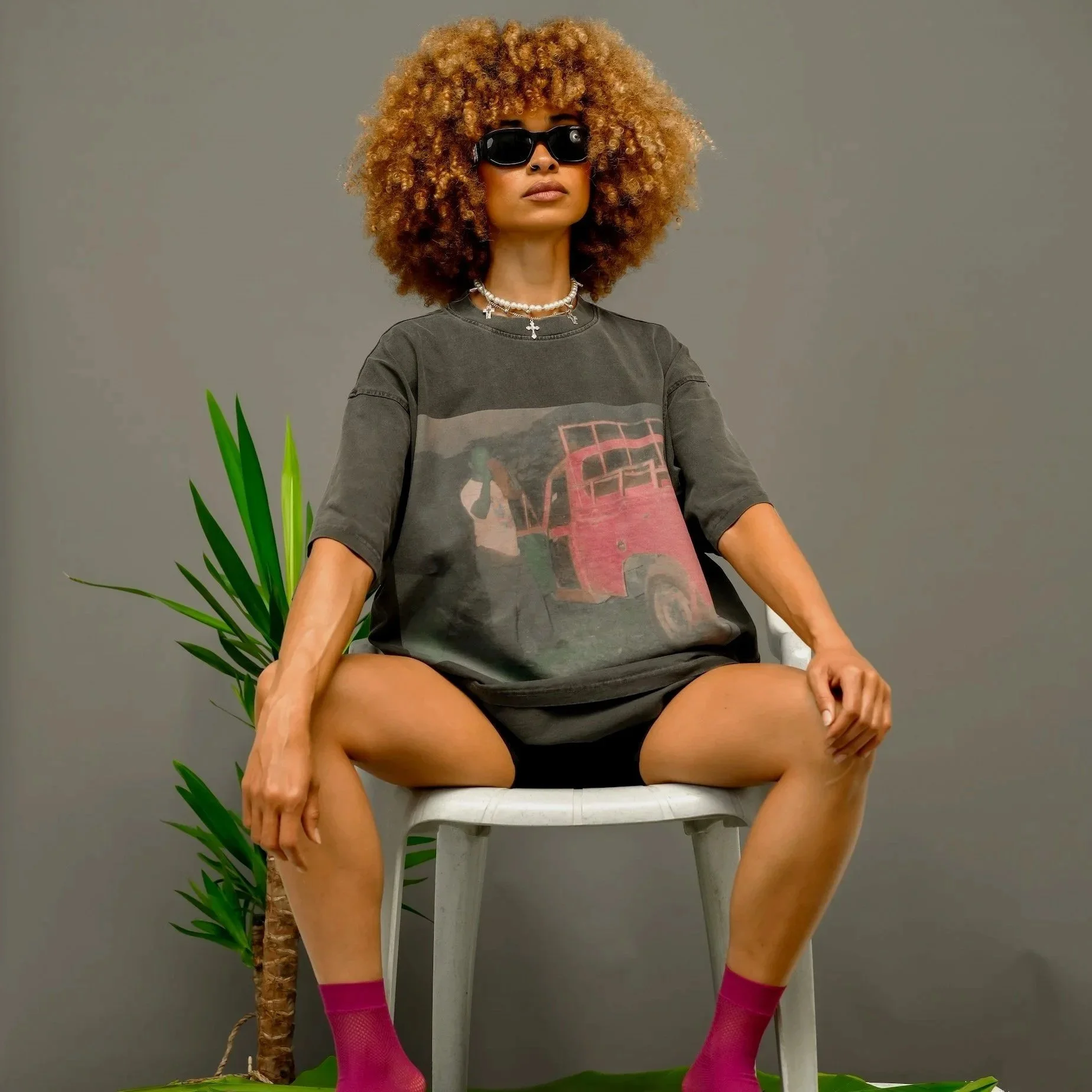 Woman sitting on a white chair with her legs spread apart, wearing a graphic T-shirt, black shorts, pink fishnet stockings, and sunglasses, with curly hair, a pearl necklace, and a neutral facial expression, positioned in front of a gray background with a green plant.