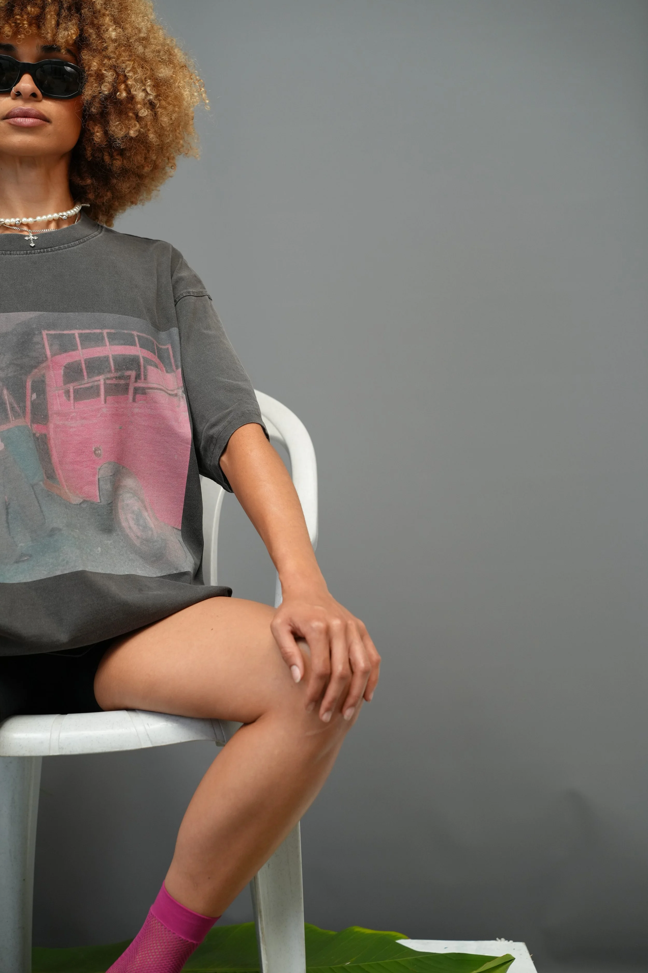 A woman with curly hair wearing sunglasses, a pearl necklace, a graphic t-shirt with a pink truck, and pink fishnet tights is sitting on a white chair, with her right leg crossed over her left knee, in front of a plain gray wall.
