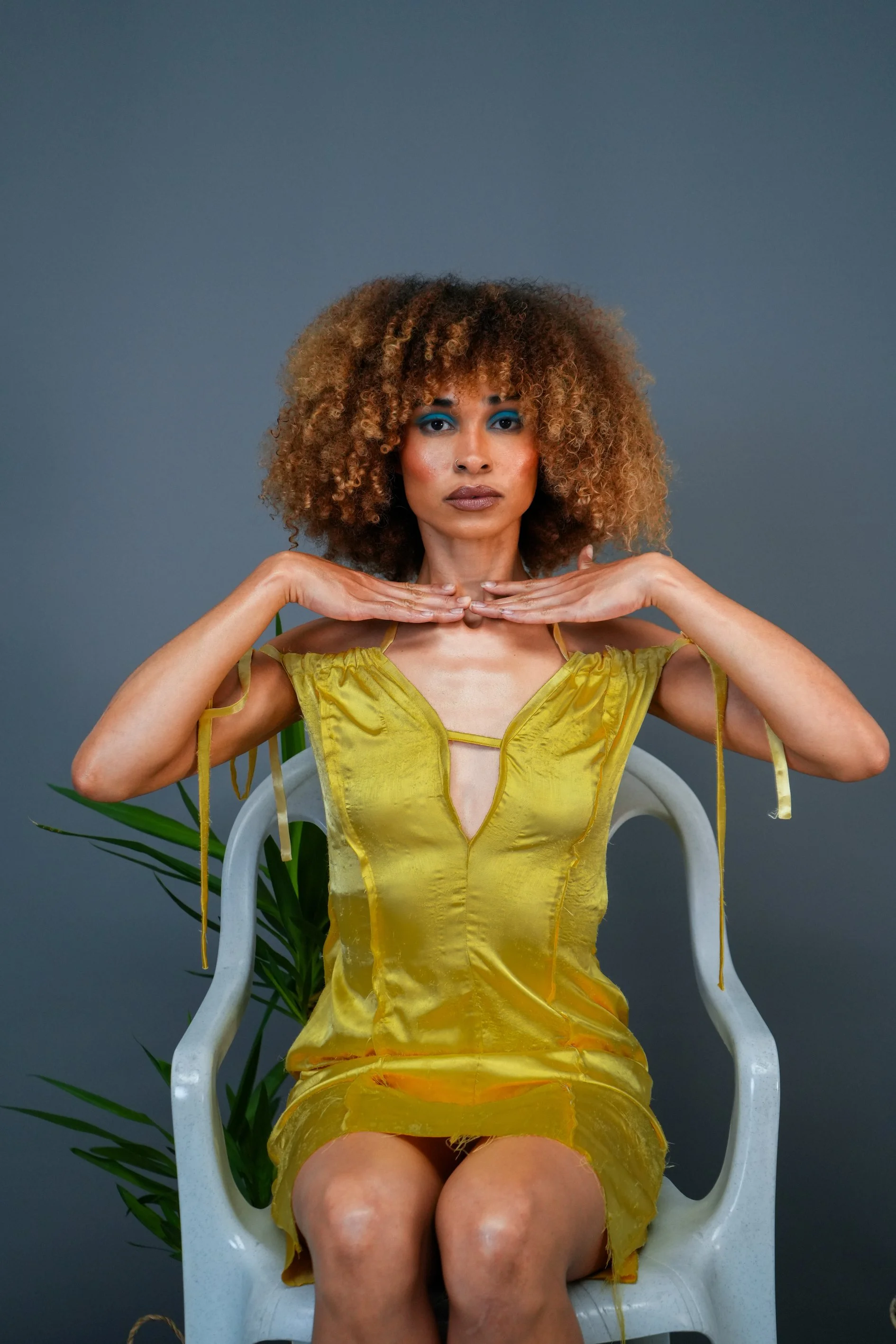 A woman with curly hair and blue eyeshadow sitting on a white chair wearing a yellow dress with a slit, with her hands under her chin against a gray background and a green plant nearby.