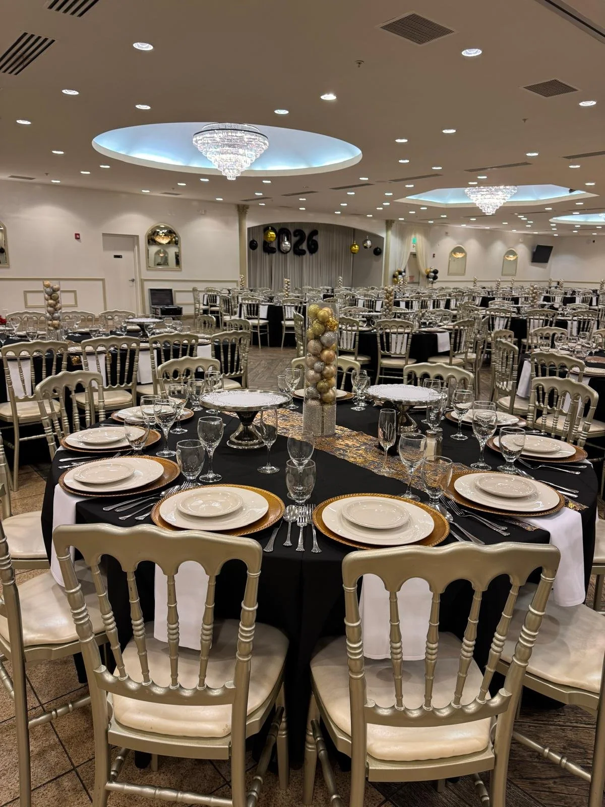 Crystal Palace Banquets decorated for a New Years Party. The room has black, white, and gold decorations, and there is a large 2026 in the background. The tables are set for dinner and have gold and silver centerpieces.
