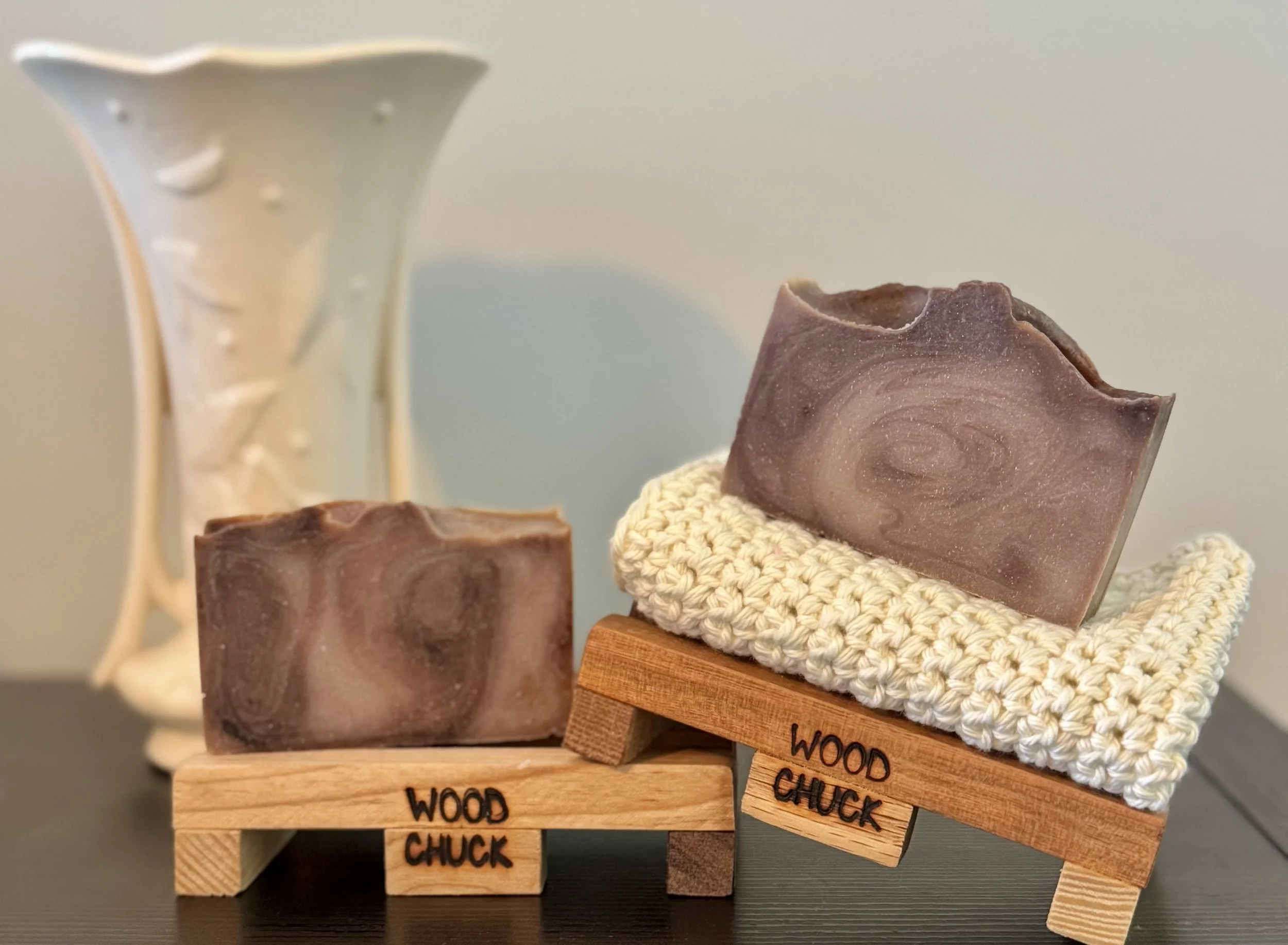 Two bars of handcrafted soap, one placed on a small wooden stand with "Wood Chuck" engraved, and the other on a knitted soap pad on a tilted wooden stand, with a white decorative vase in the background.