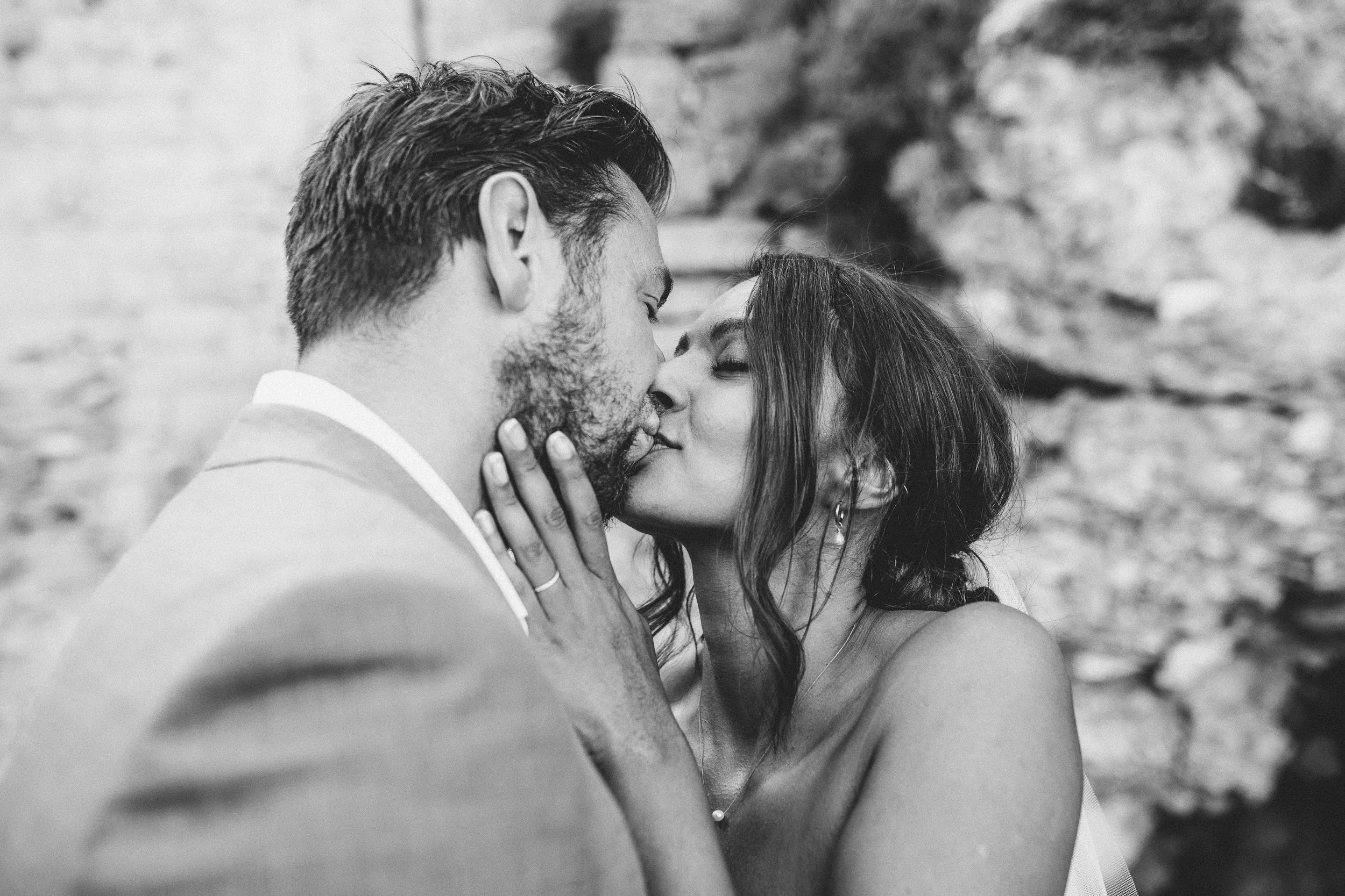 A black-and-white photo of a couple in an intimate moment, their faces close as they touch noses and hold each other's faces, with a blurred natural background.