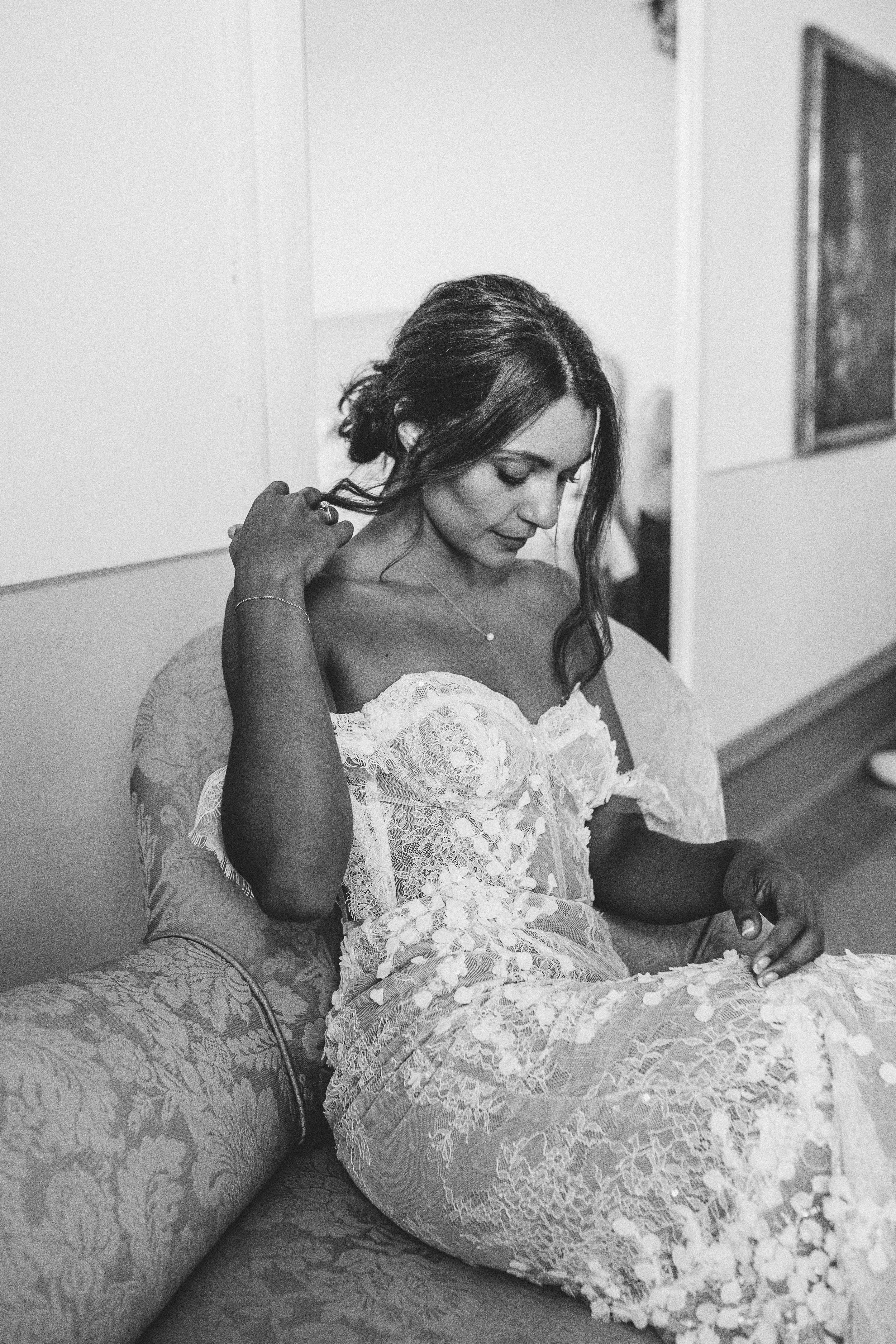 A bride in a lace dress seated on a patterned armchair, looking down and touching her hair, with a mirror visible in the background.