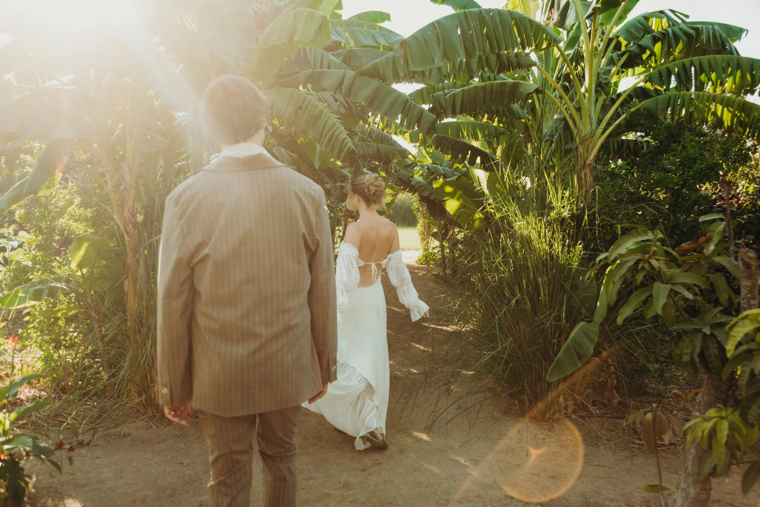 People walking through a lush green tropical garden or forest, with sunlight filtering through the leaves, including a woman in a white off-shoulder dress and a man in a striped suit.