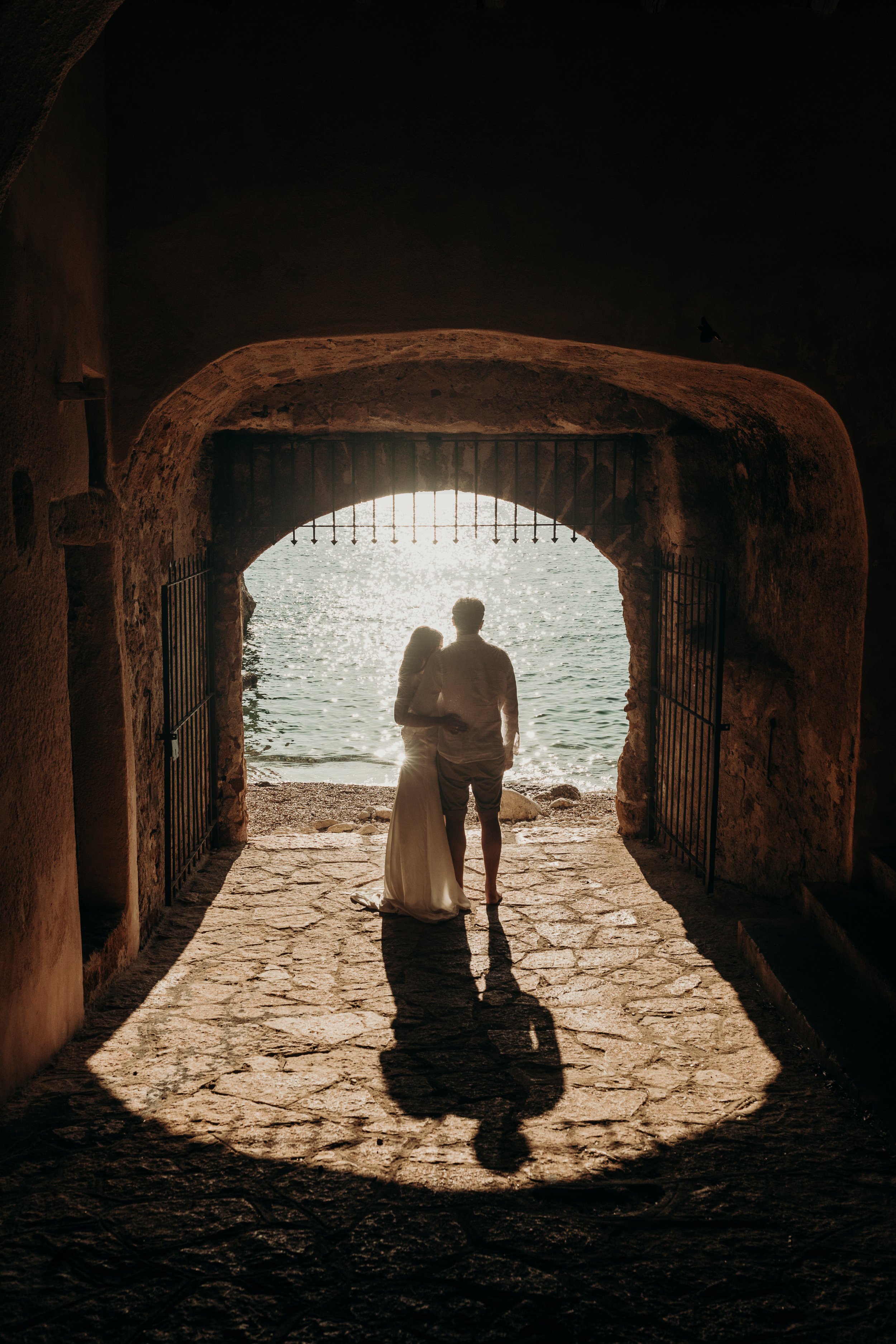 A couple standing together under an archway overlooking the water at sunset, with their shadows cast on the stone ground.