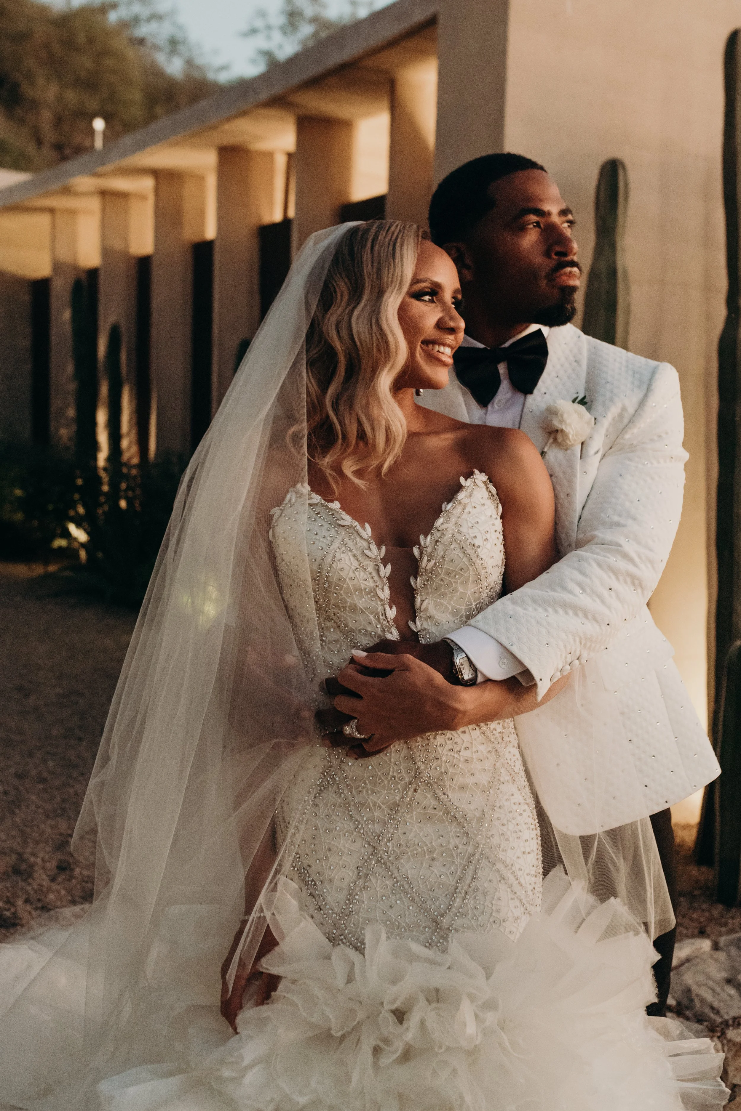 A newlywed couple, the bride in a white wedding gown with intricate beading and lace, and the groom in a white tuxedo with a black bow tie, embracing each other during their wedding celebration with warm evening lighting.
