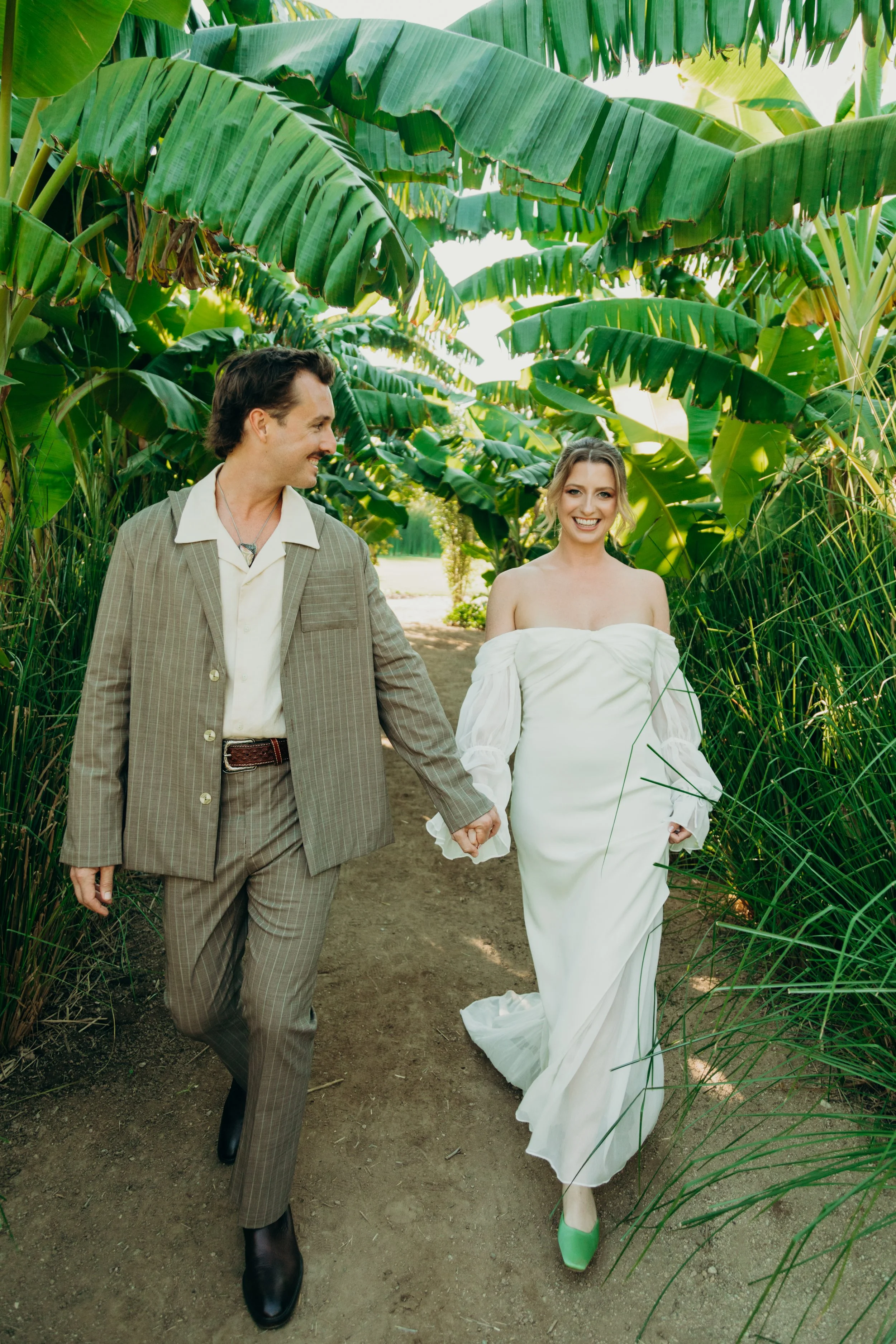 A couple is walking through a lush, green banana plant garden, holding hands. The man is dressed in a tan checked suit with a white shirt, and the woman is wearing a white off-the-shoulder dress with green shoes. They are smiling and looking at each 