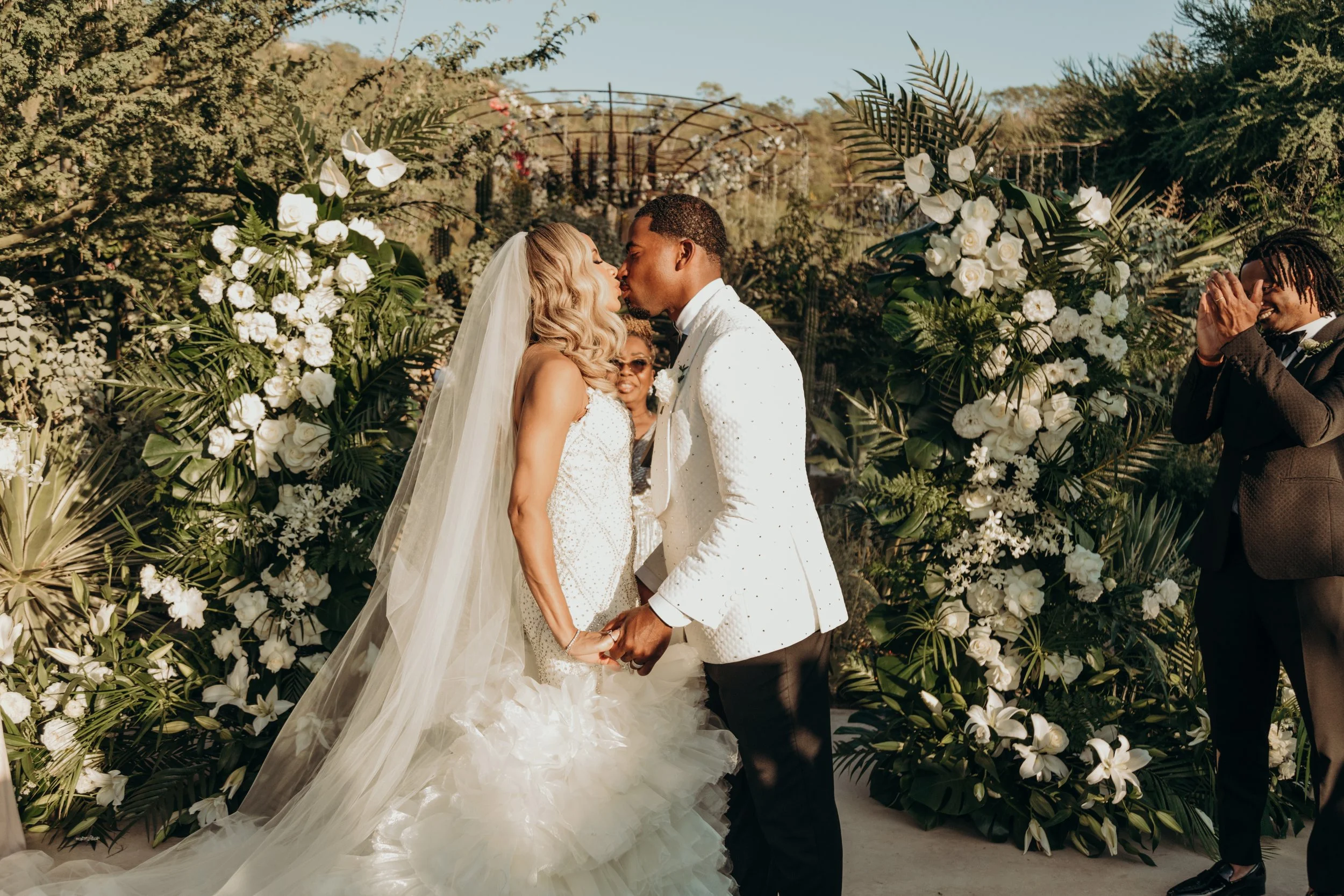 A wedding ceremony with a bride and groom kissing, surrounded by floral arrangements, with a person on the right reacting emotionally and an officiant or witness in the background.