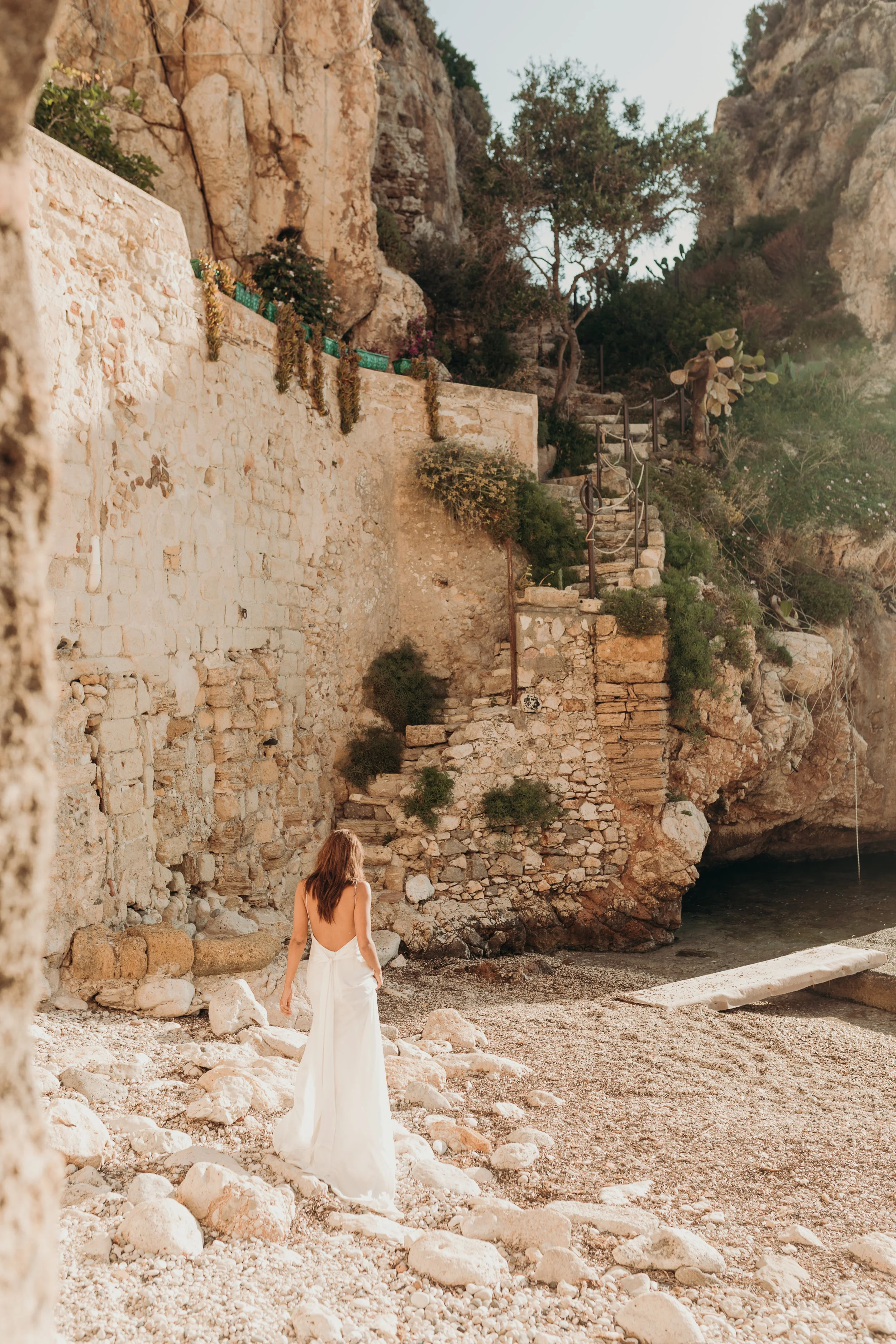 Woman in a white dress walking on a rocky beach with stone stairs and a stone wall built into the cliffside, surrounded by trees and cacti.