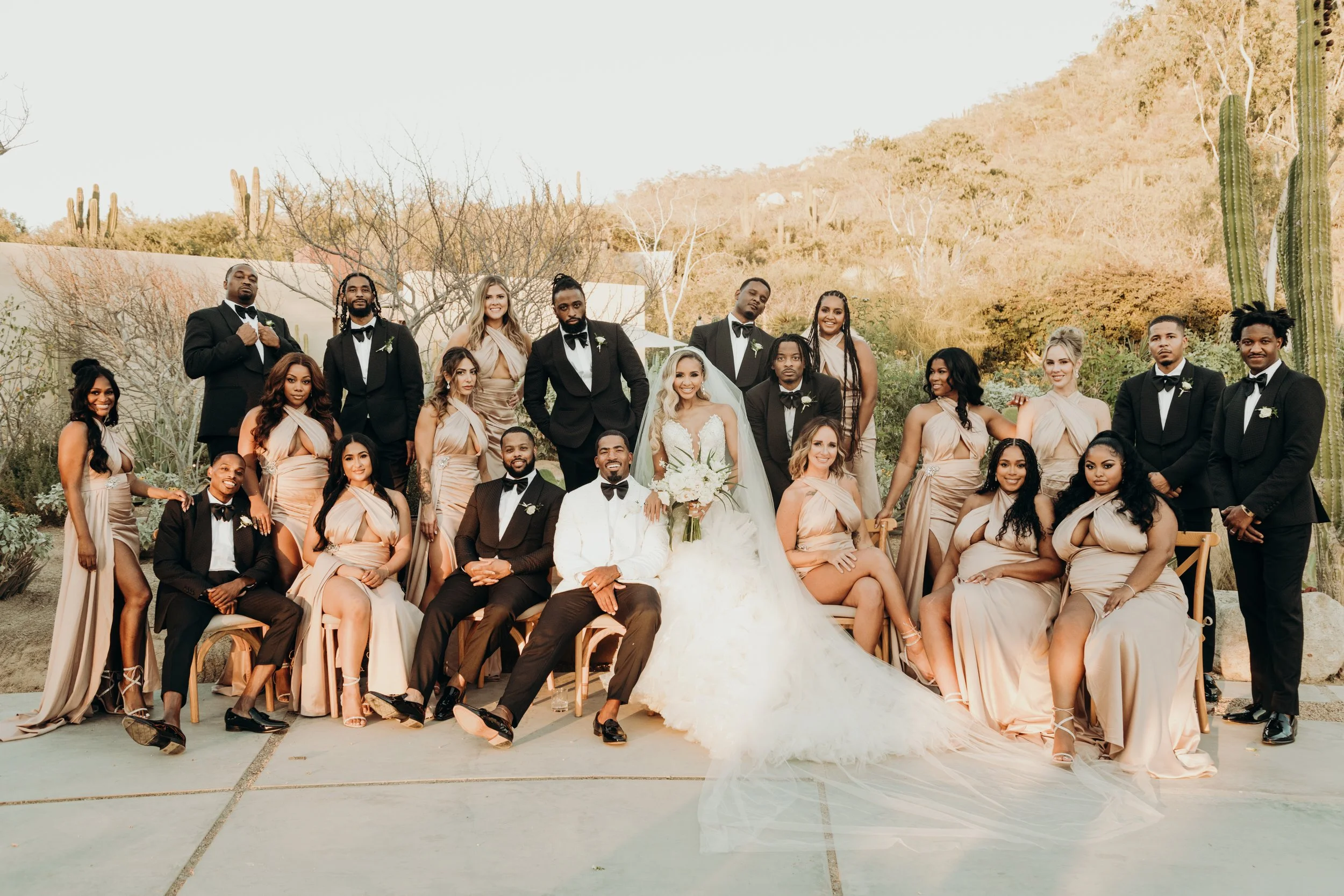 A wedding party outdoors with roughly 20 people, including the bride in a white gown and veil, and groom in a black tuxedo, surrounded by bridesmaids in beige dresses and groomsmen in black tuxedos, with desert plants and cacti in the background.