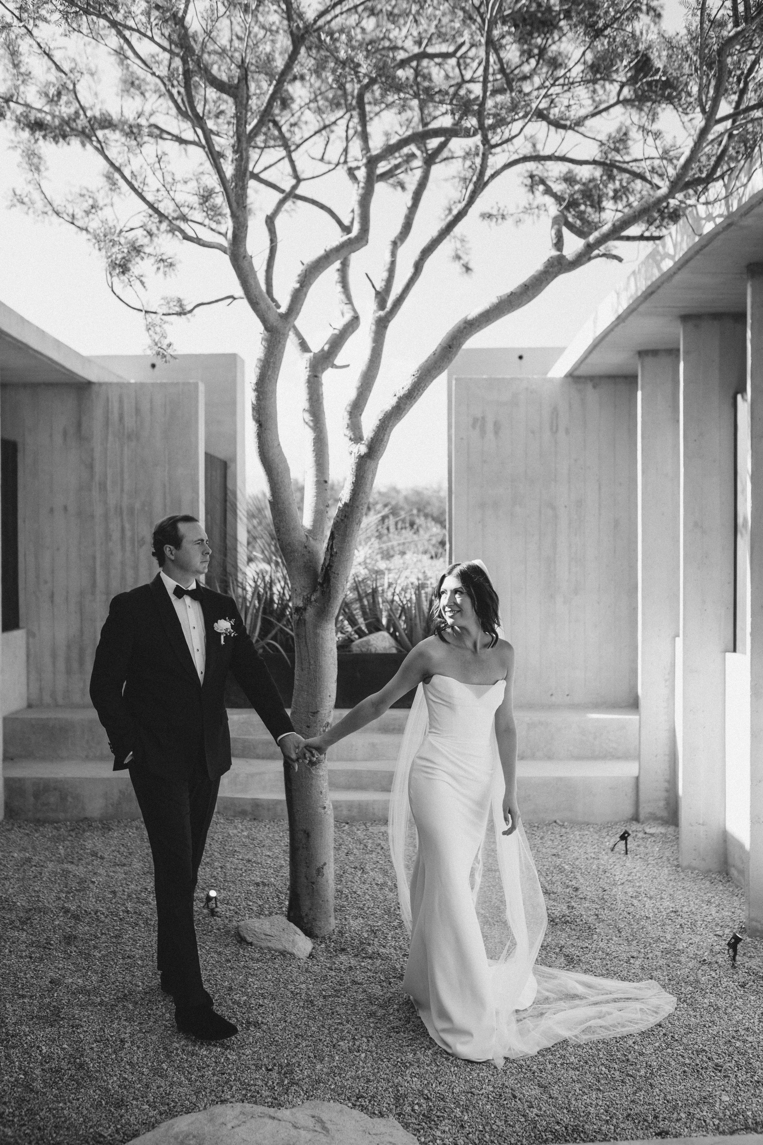 A black-and-white photo of a man in a tuxedo holding hands with a woman in a strapless wedding dress, walking outdoors near a modern building with columns and a tree in the foreground.