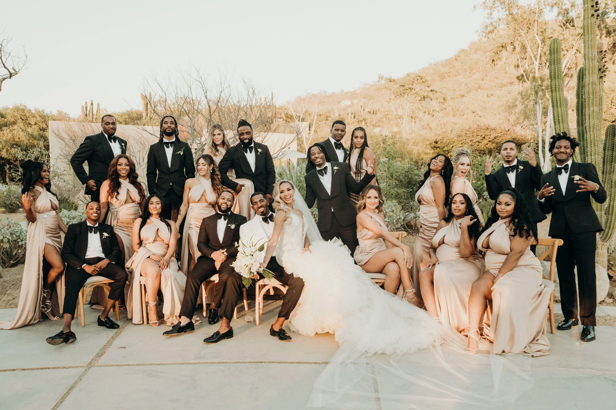 A group of people at a wedding, with bridesmaids in beige dresses and groomsmen in black tuxedos, seated and standing outdoors in a desert-like landscape with cacti and mountains in the background.