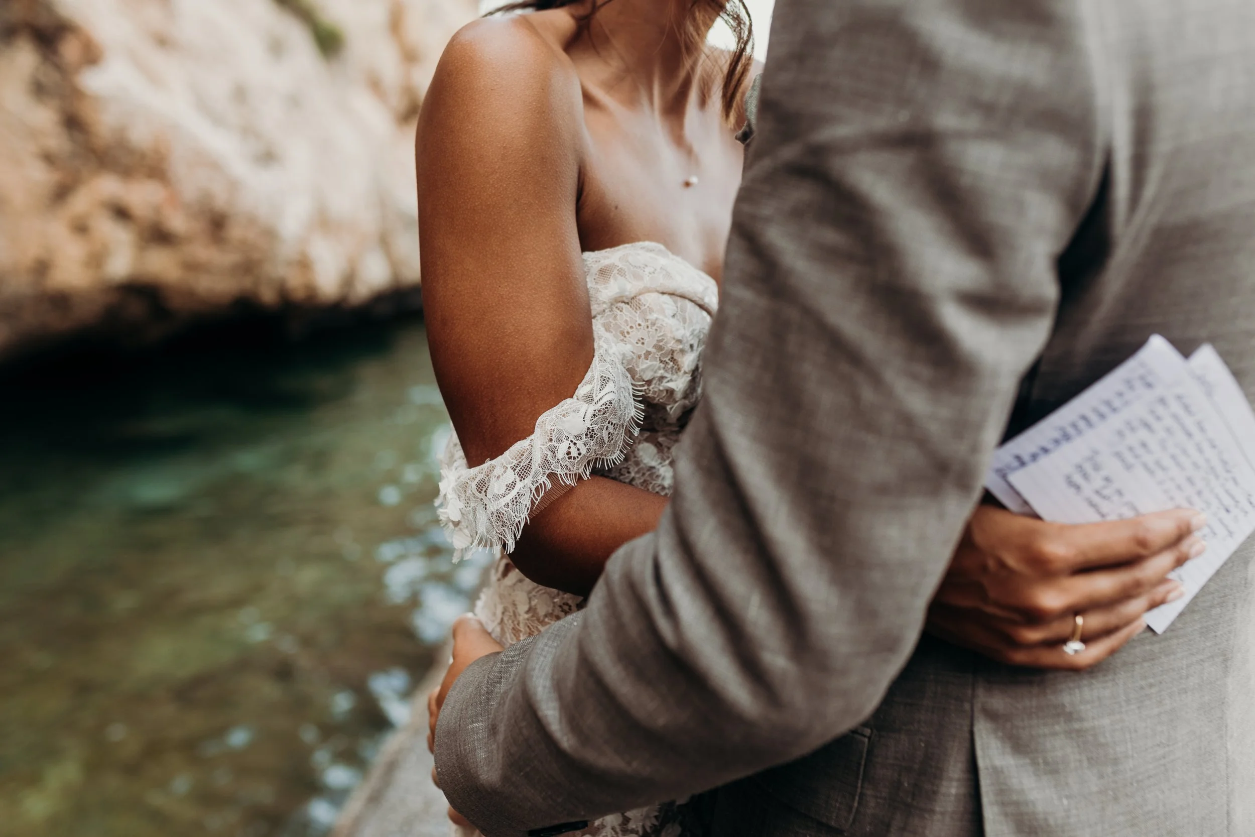 Close-up of a woman in a white lace off-shoulder dress embracing a man in a gray suit by a river or stream, holding a piece of paper.