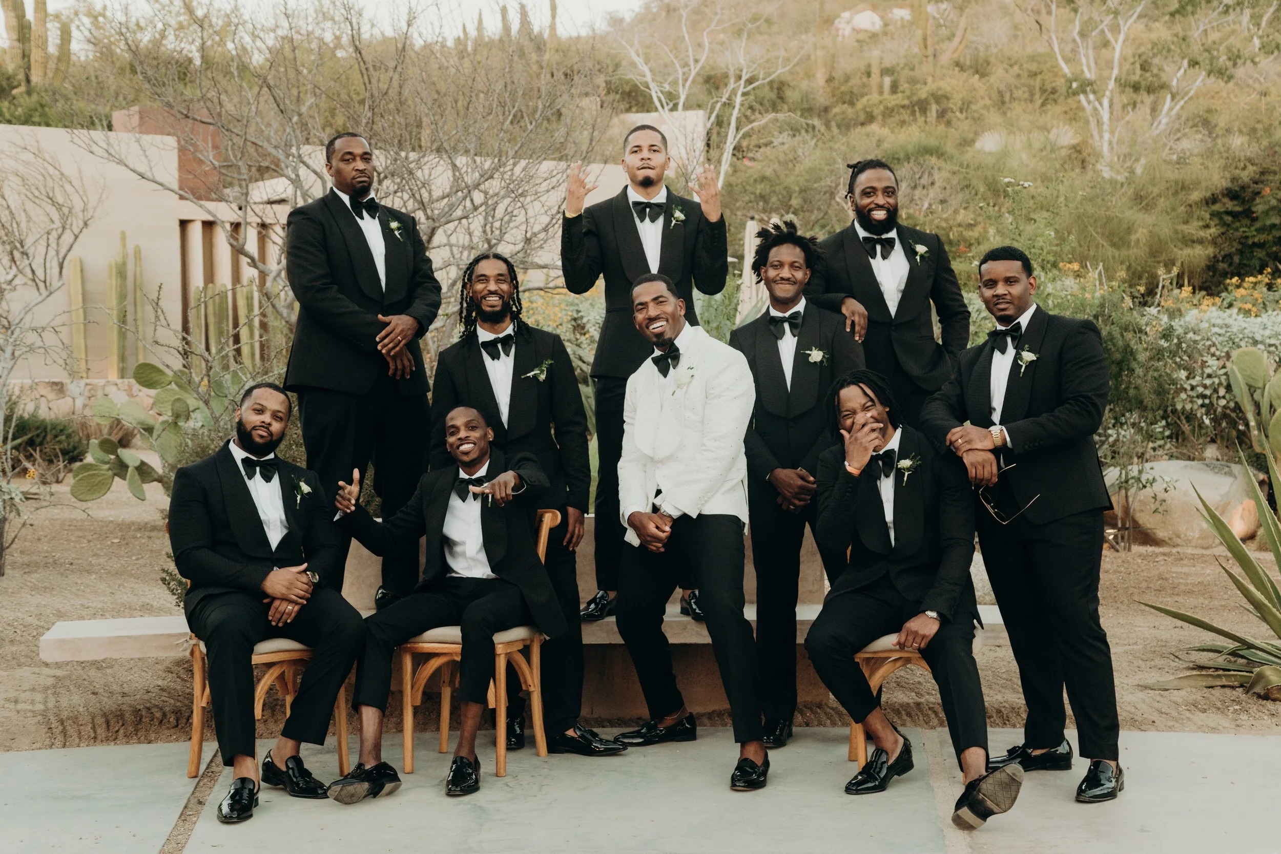A group of nine men dressed in formal black tuxedos with bow ties, posing outdoors during daytime. The setting includes desert plants like cacti and trees with some leaves, with a building and hillside in the background. They are smiling, laughing, a