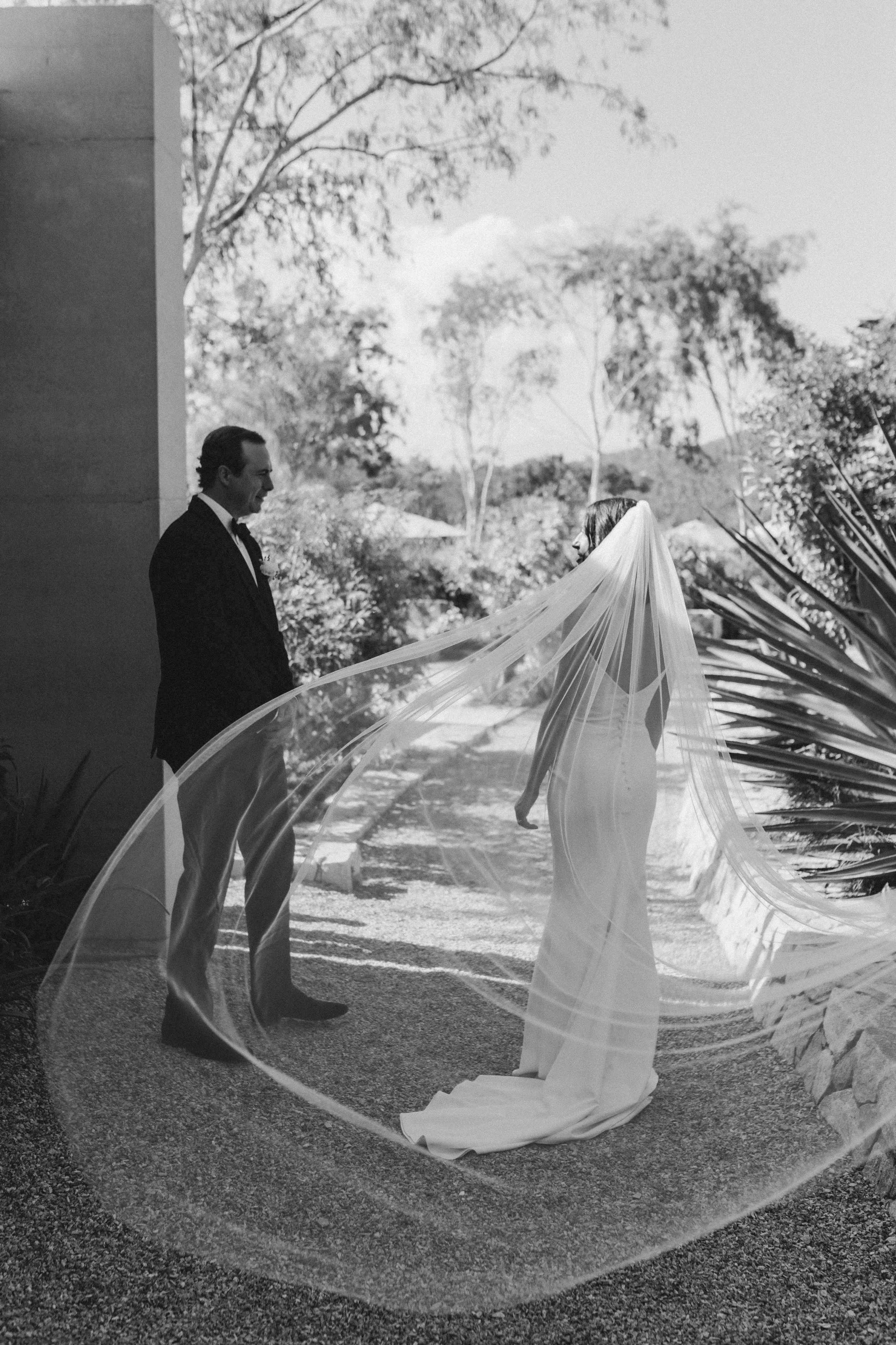 A bride and groom are standing outside facing each other, with the bride's veil flowing and the groom wearing a suit, in a garden setting with trees and plants in the background.