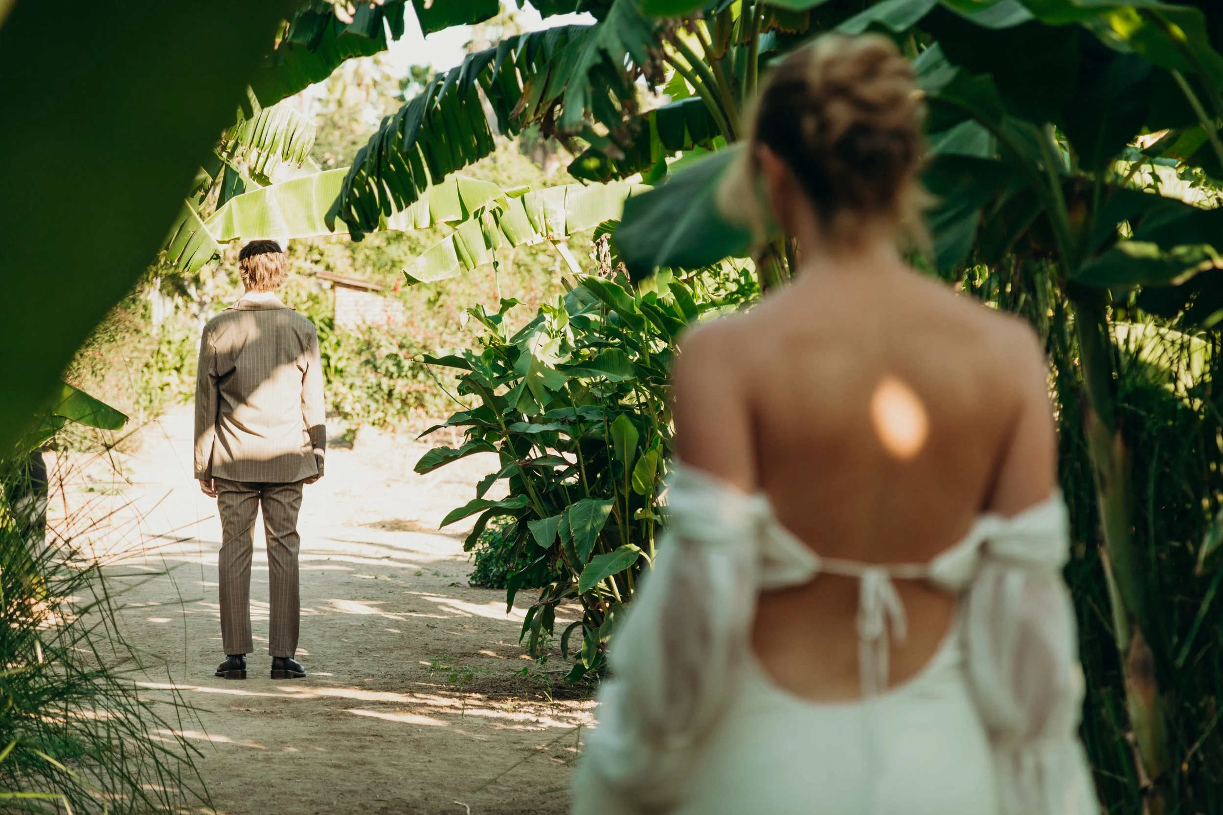 A woman with her back to the camera wearing a cream-colored top, standing among large green banana leaves. A man in a checkered suit is walking away on a dirt path ahead of her, surrounded by lush greenery and plants.