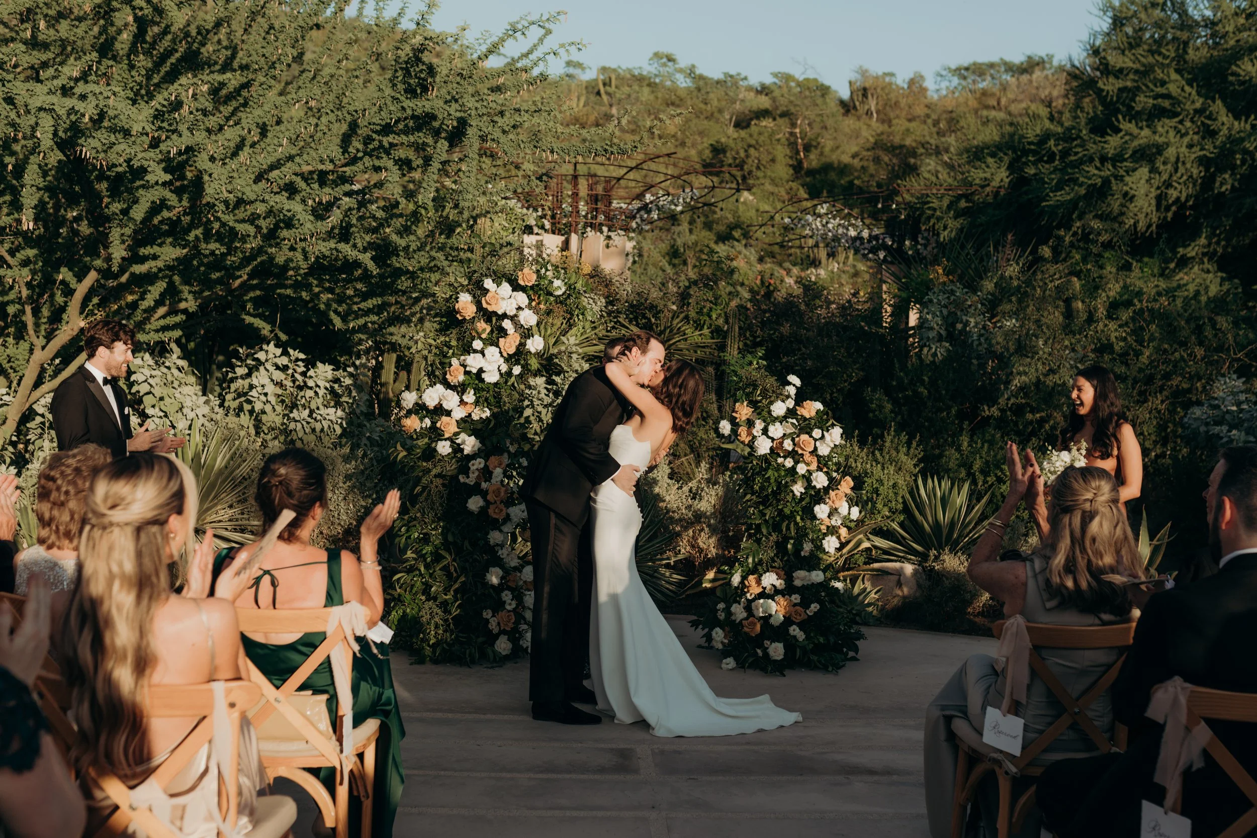 A bride and groom kiss during an outdoor wedding ceremony with guests seated and clapping, surrounded by lush green trees and floral arrangements.