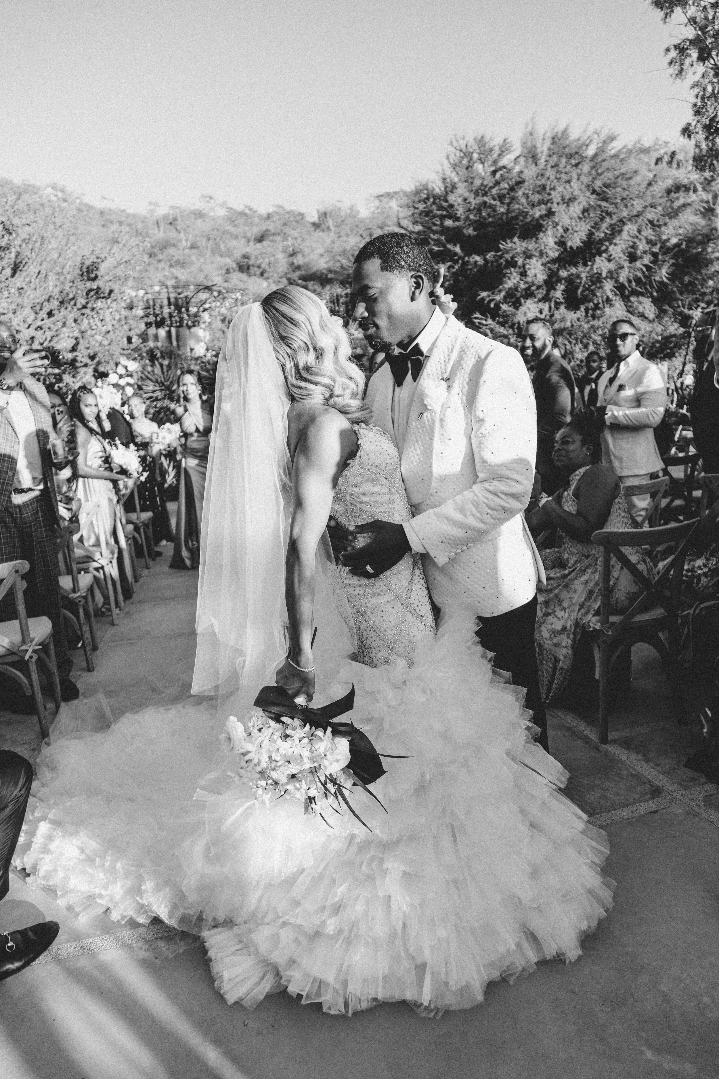 A black and white photo of a wedding ceremony outdoors. The bride and groom are standing close together, holding hands and leaning in for a kiss. The bride is wearing a voluminous wedding dress with a long train and a veil, and the groom is in a tuxe