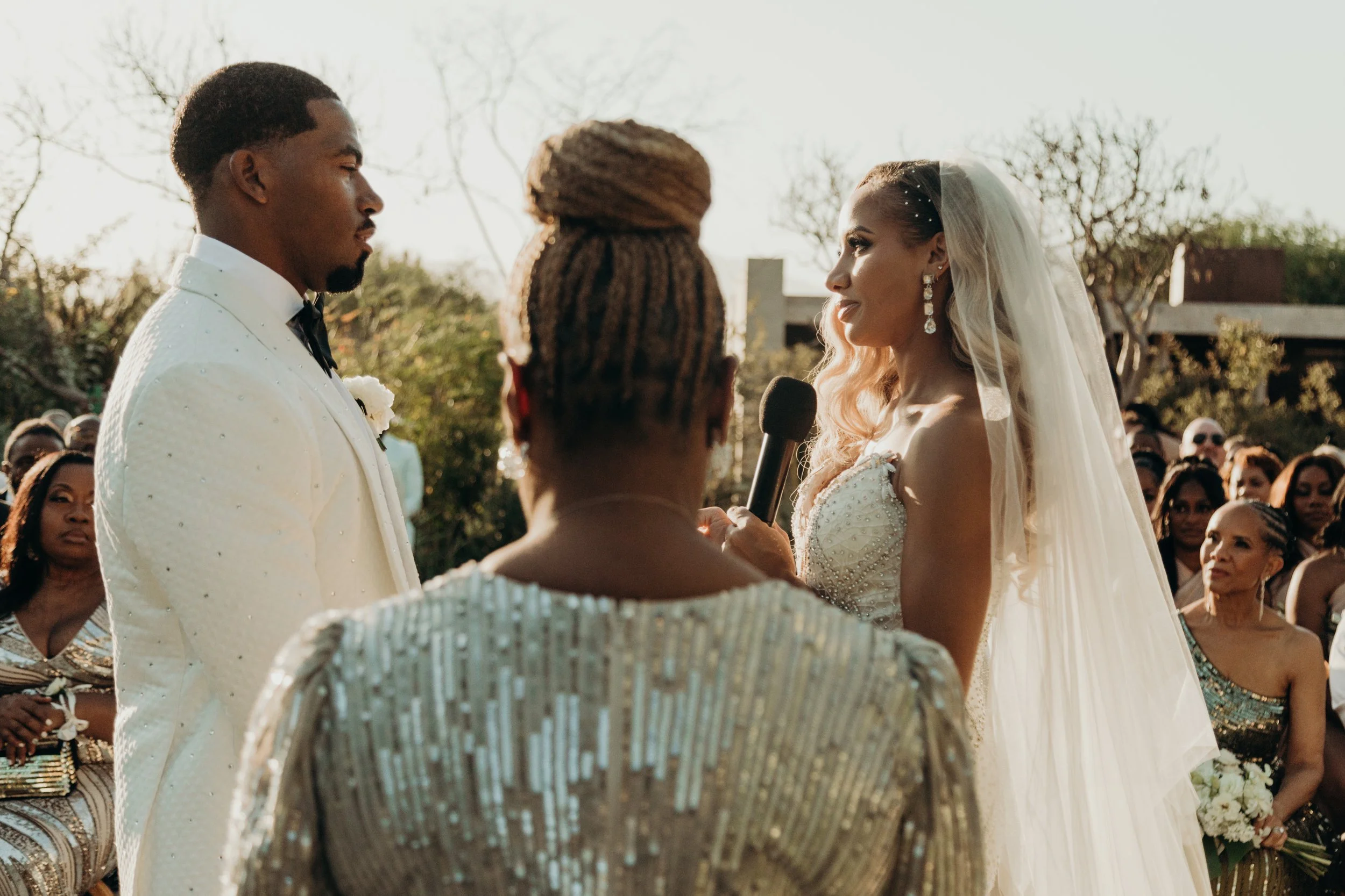 A bride and groom exchange vows during their outdoor wedding ceremony, surrounded by guests, on a sunny day.
